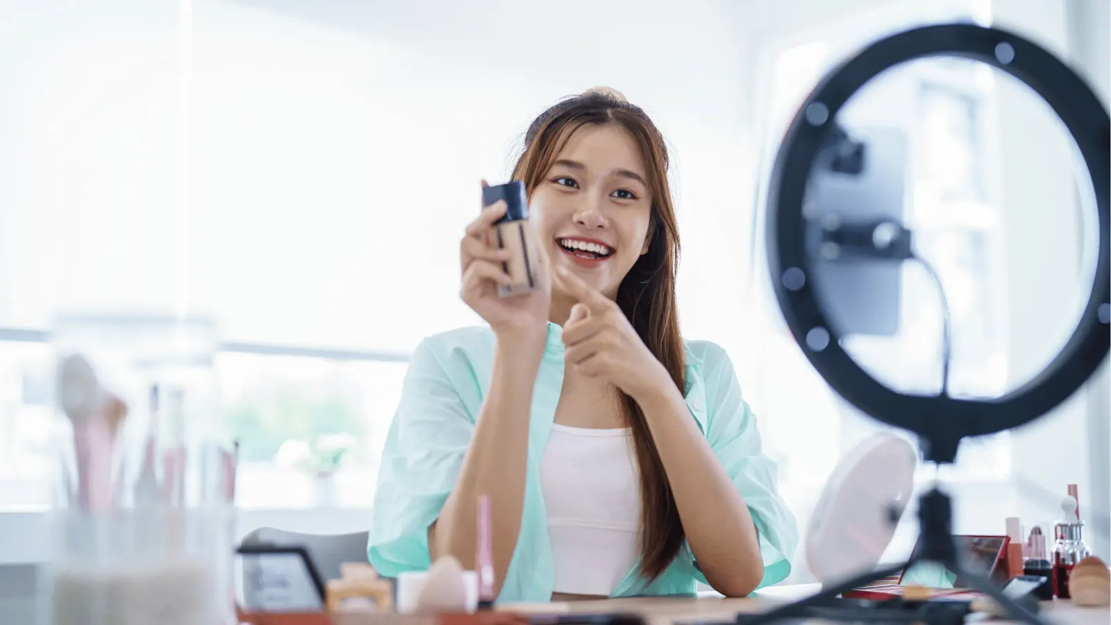 Young woman vlogger holding foundation bottle while filming with ring light