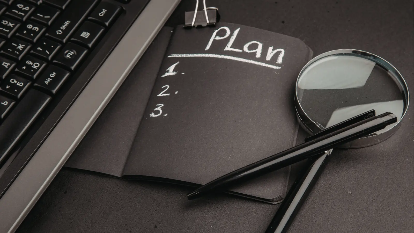 Black notebook labeled 'Plan' with magnifying glass, pens, and laptop keyboard