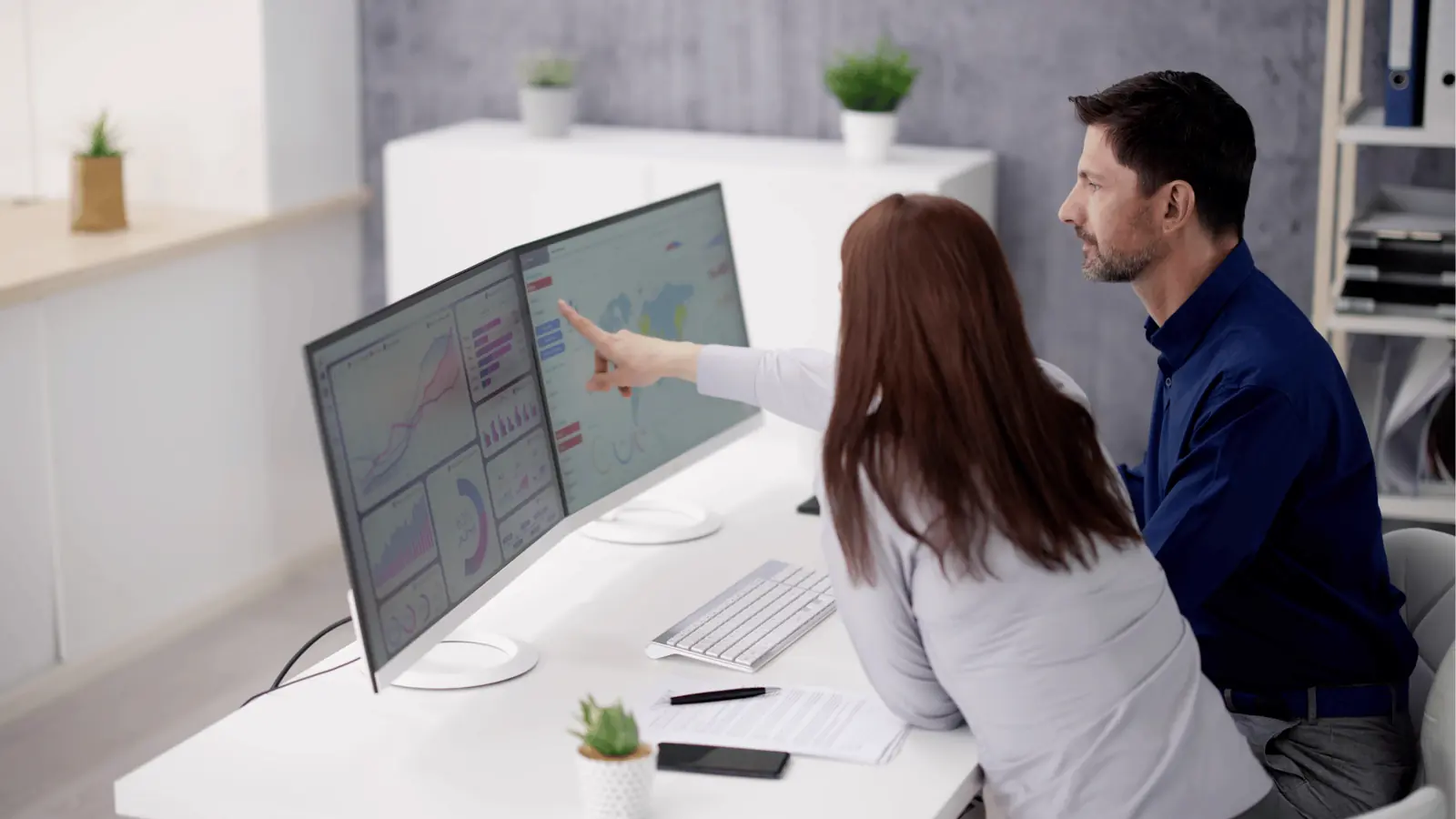 Two colleagues pointing at dual monitors showing charts and maps in office for AI-assisted workflow