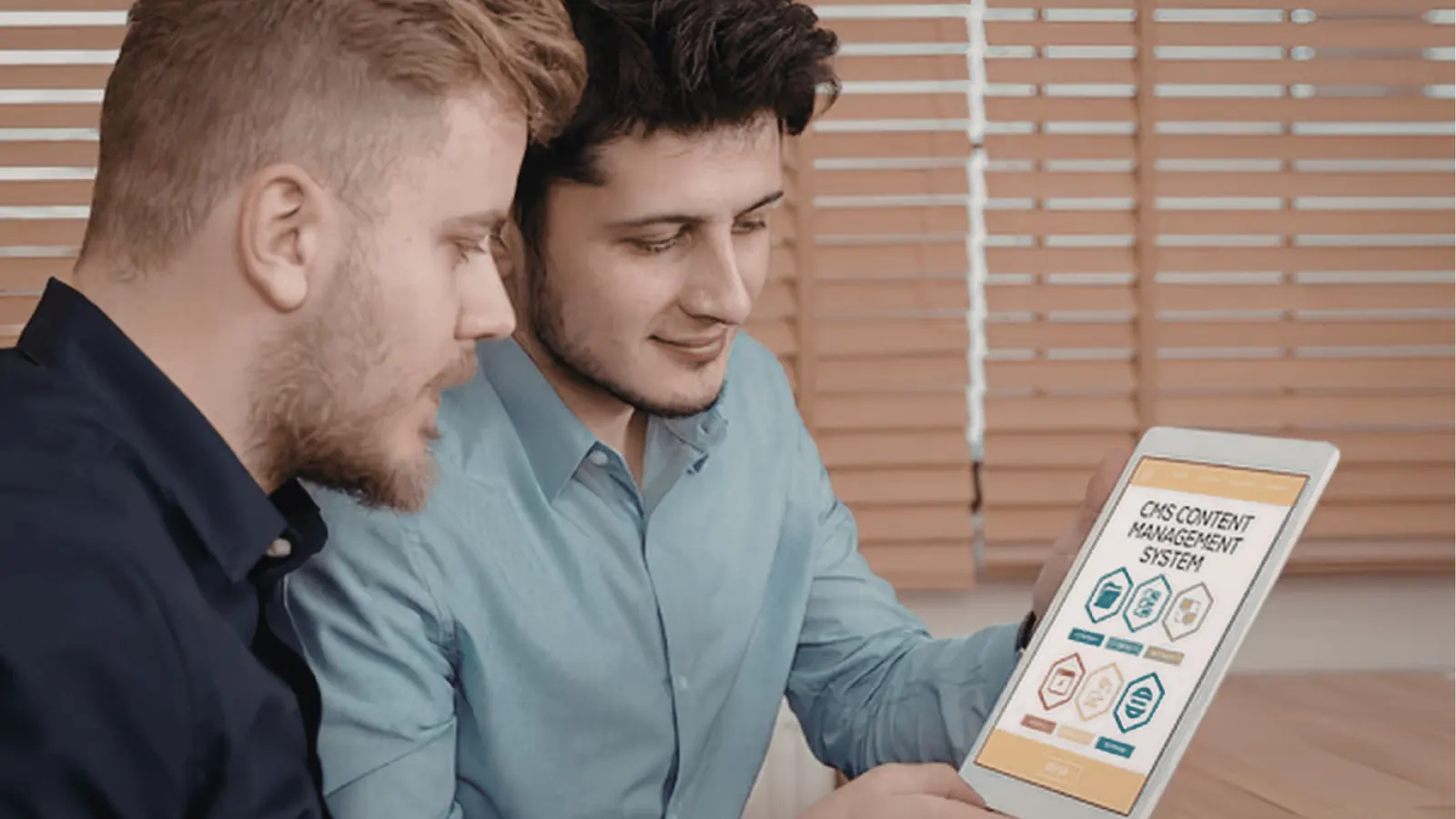 Two colleagues sitting by blinds looking at a tablet showing a CMS interface