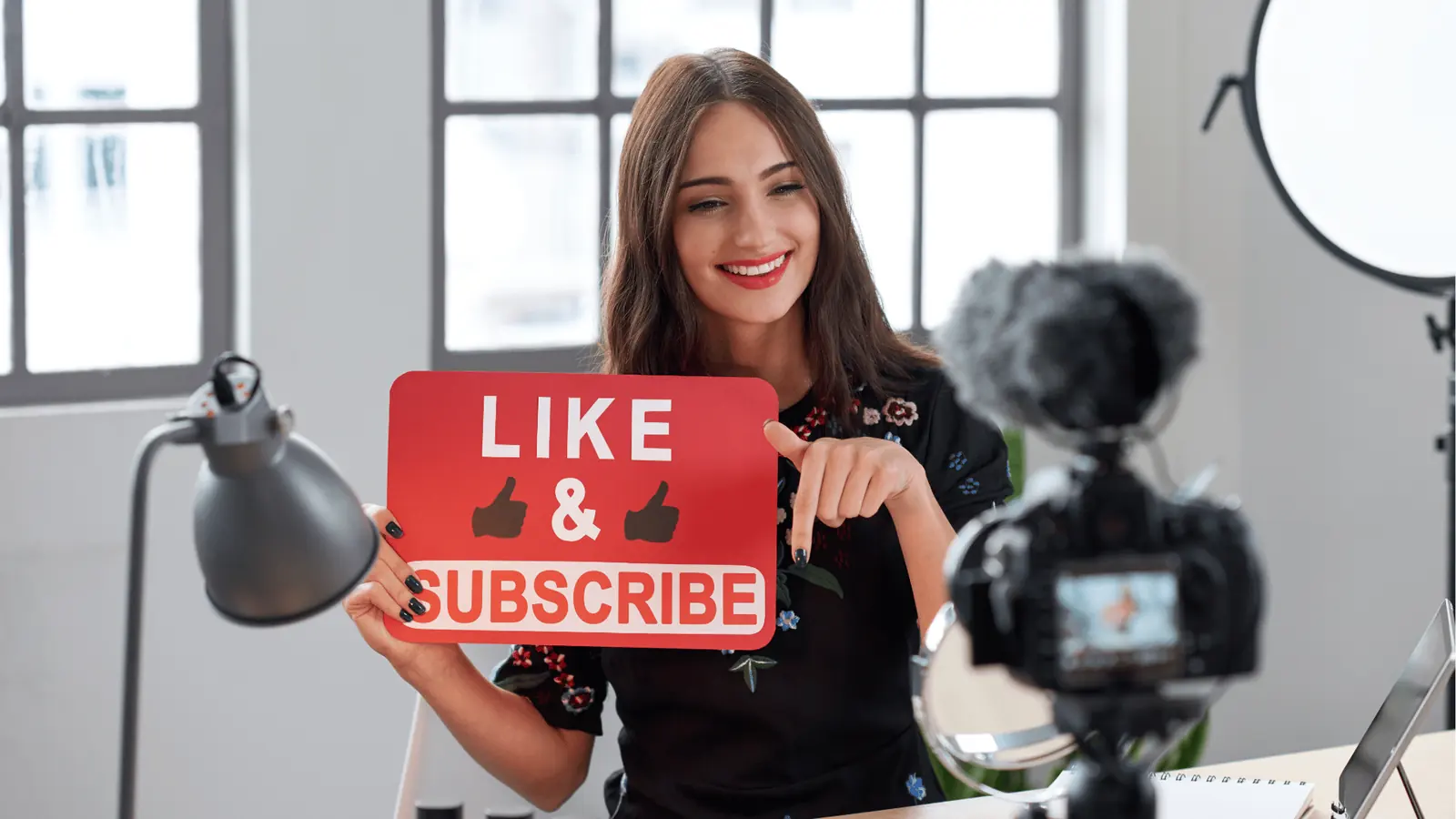 Smiling female content creator holding 'Like & Subscribe' sign in front of camera for scheduling