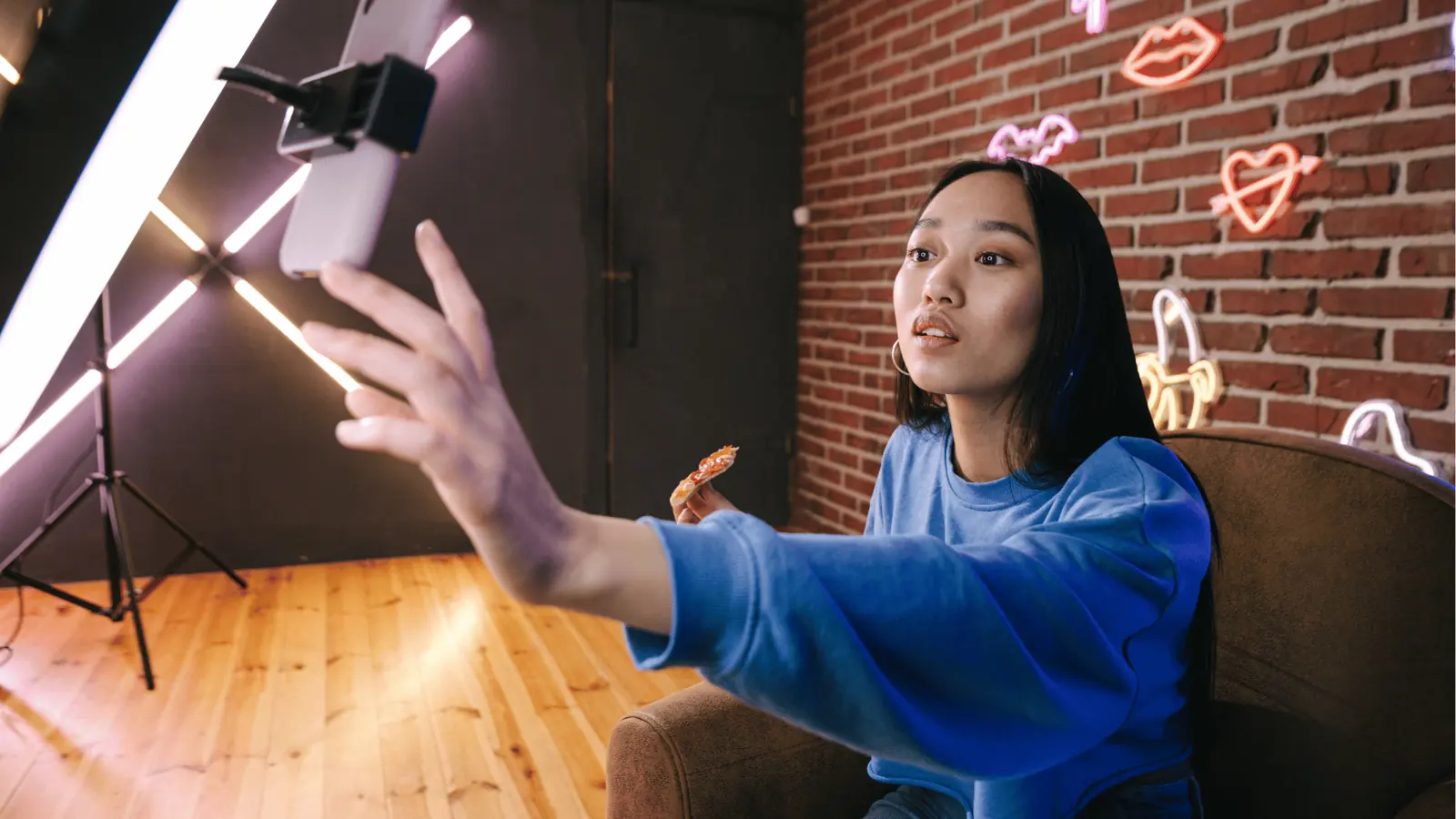 Young woman in blue sweatshirt reaching for phone while eating pizza slice