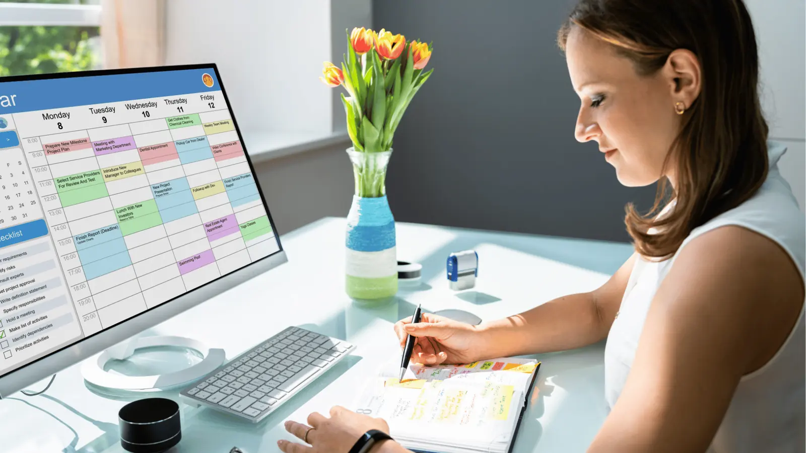 Woman writing in a planner at desk beside computer showing weekly calendar