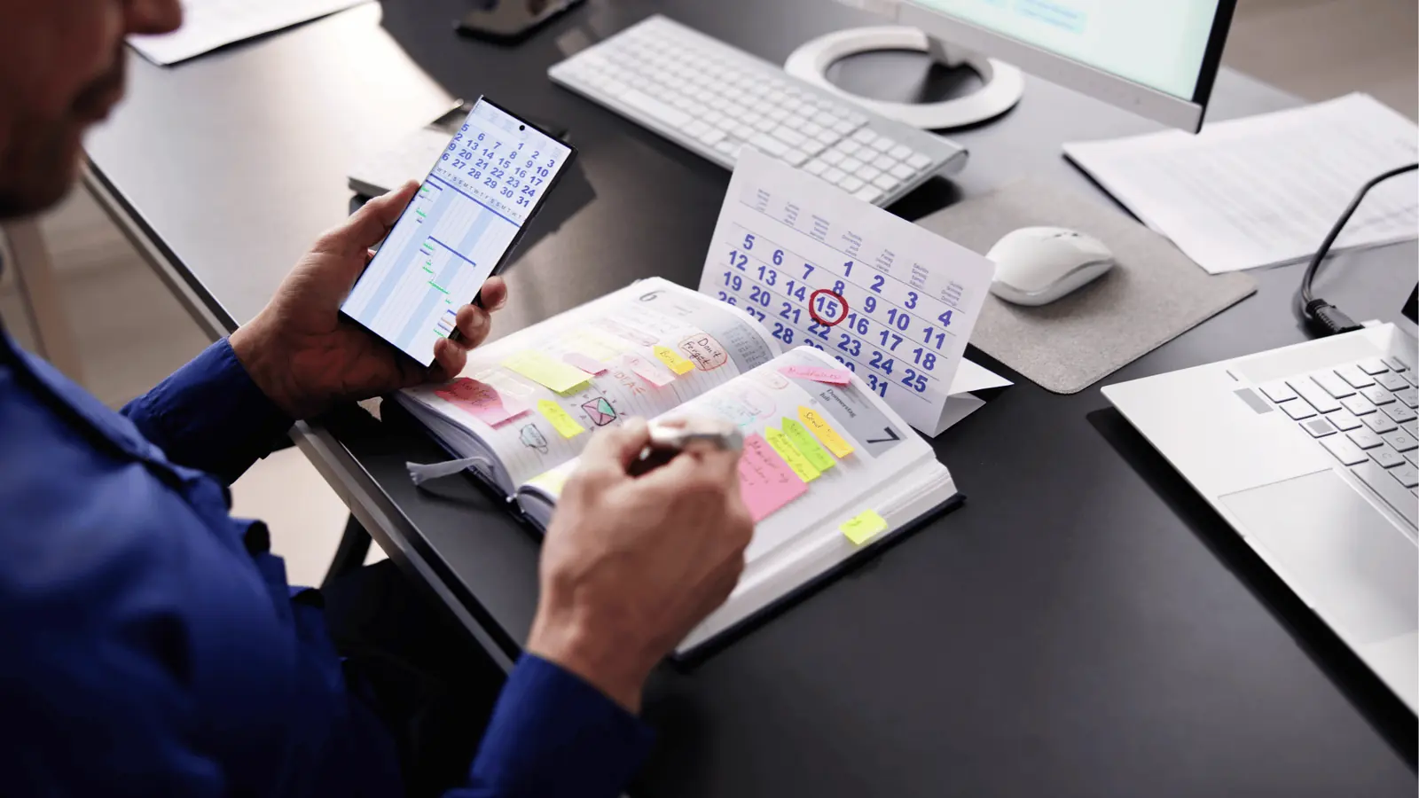 Person writing in a sticky-note filled planner beside phone and desk calendar