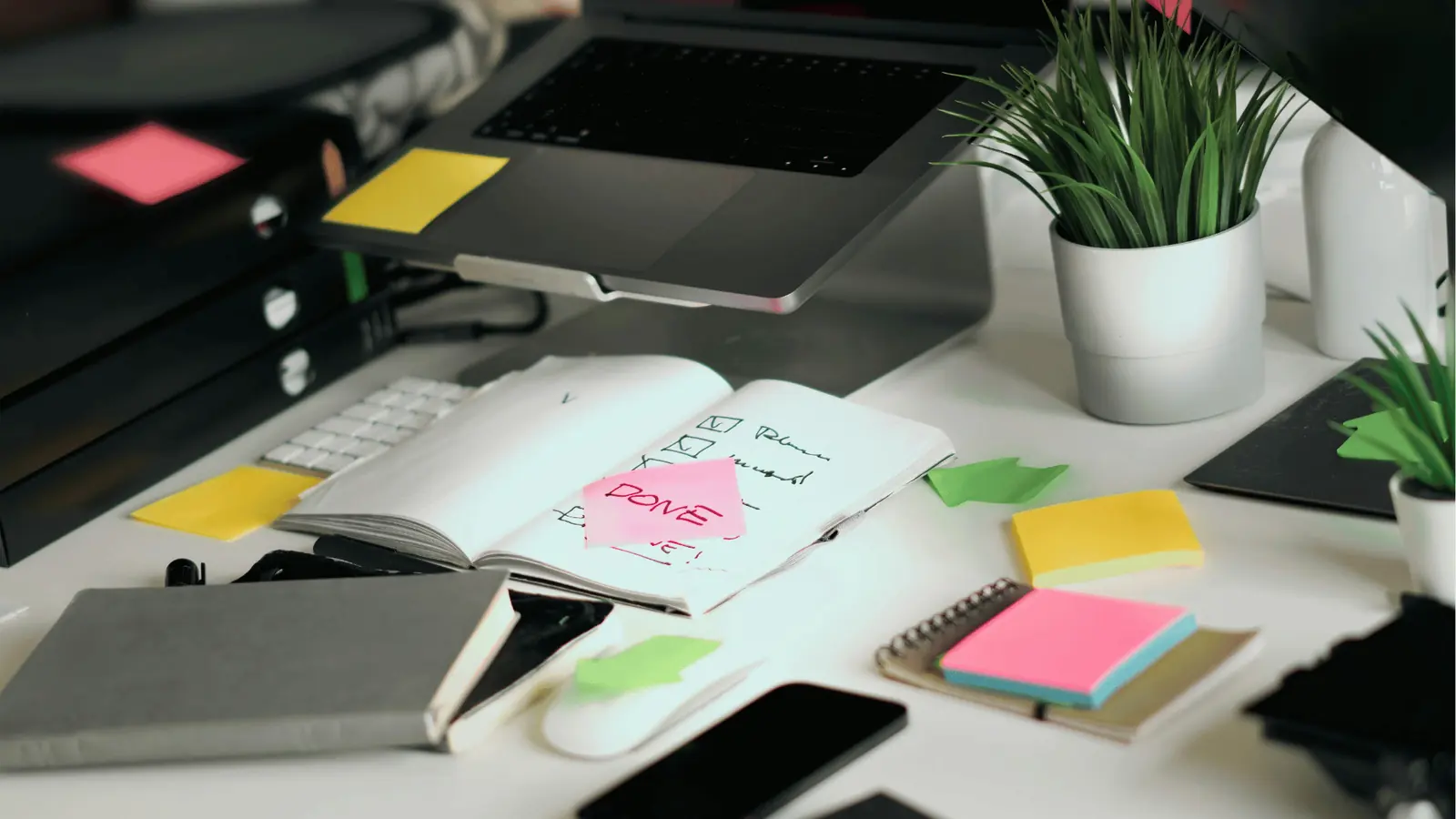 Cluttered white desk with laptop, notebooks, sticky notes and potted plant