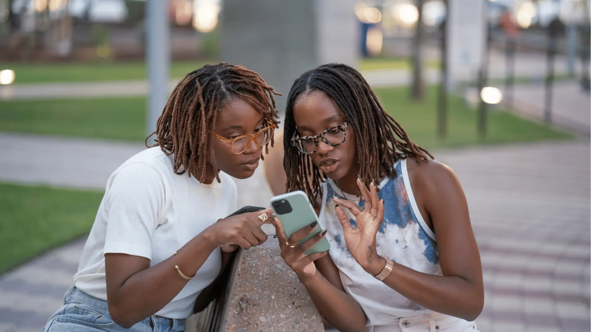 Two women with dreadlocks and glasses looking at smartphone together outdoors