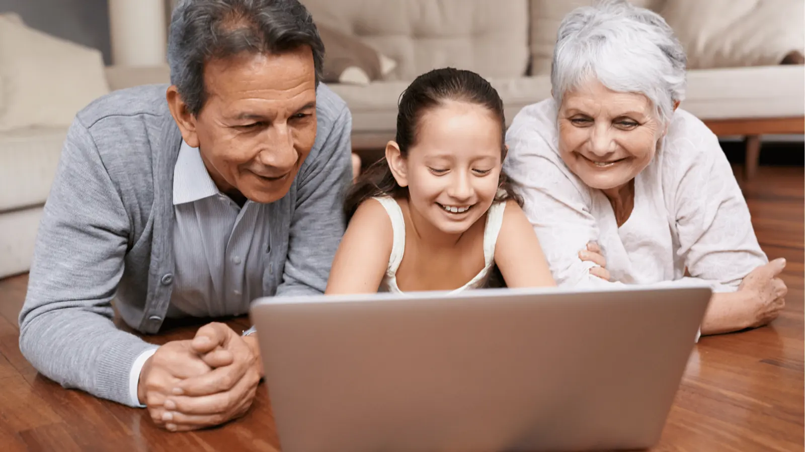 Grandparents and granddaughter lying on floor smiling while looking at laptop screen