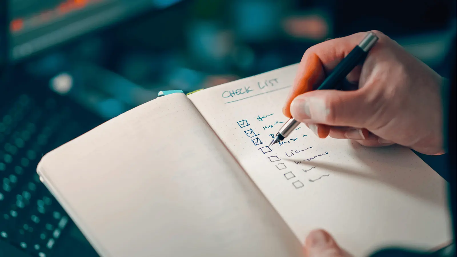 Hand marking items on a checklist in a dotted notebook near a keyboard