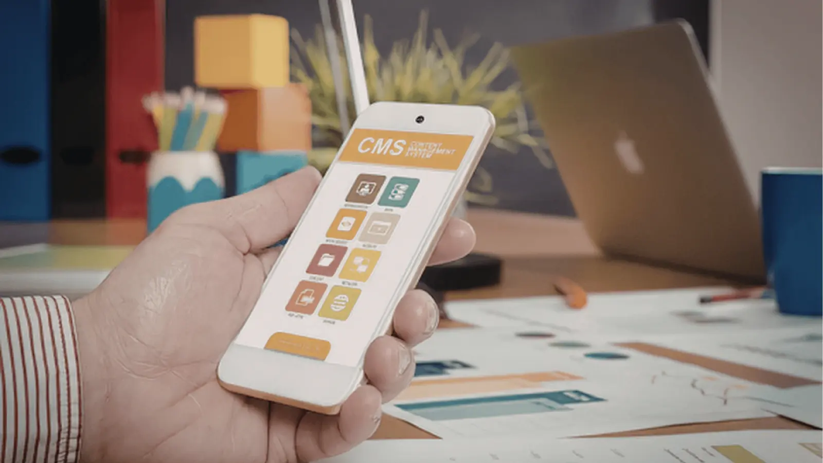 Hand holding smartphone showing CMS app UI over desk with laptop for brand management