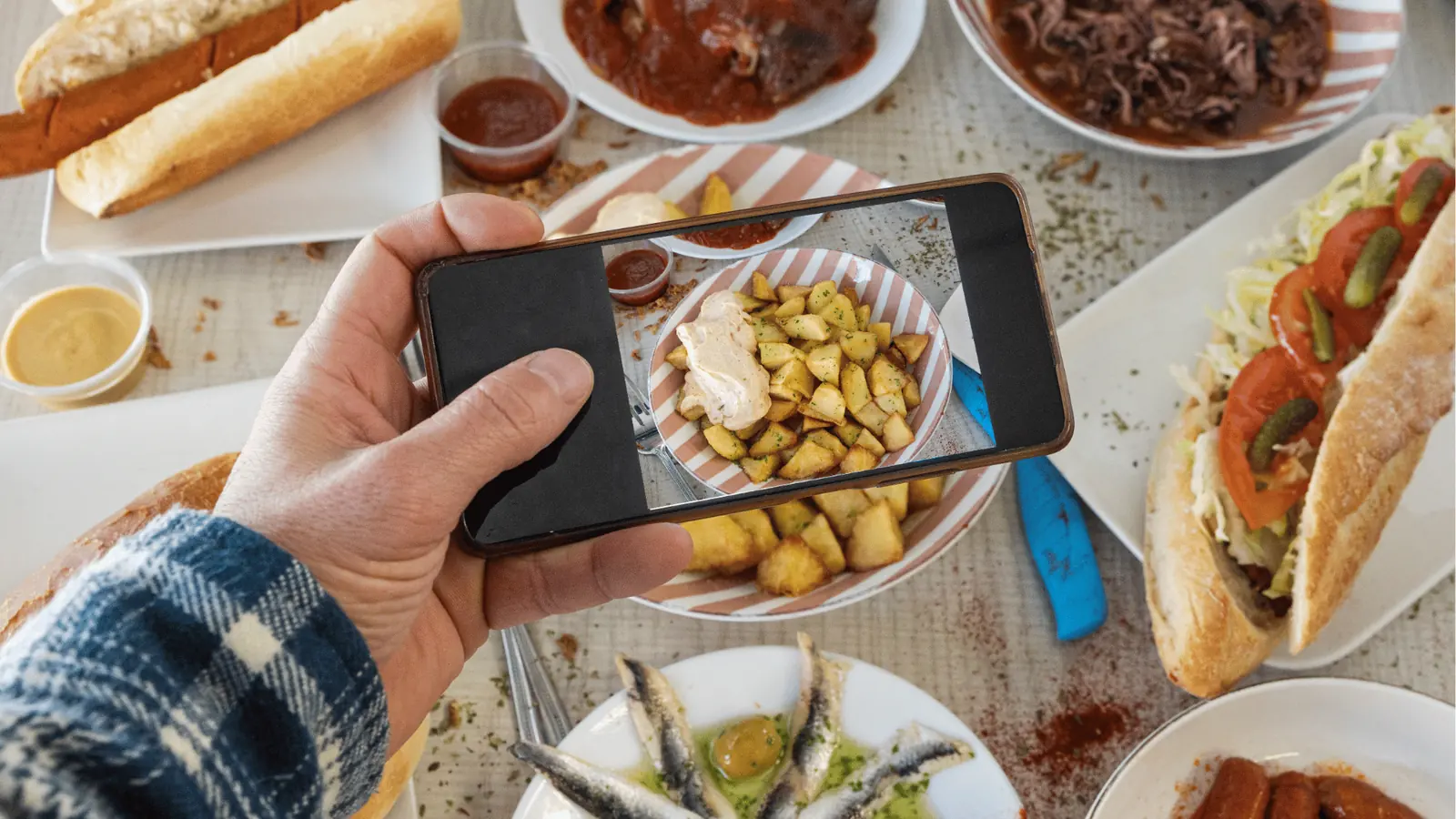 Hand holding phone photographing bowl of roasted potatoes among sandwiches