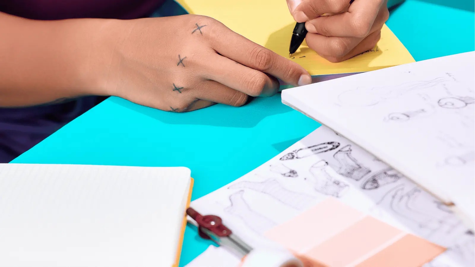 Close-up of a hand sketching on yellow paper beside notebooks and fashion sketches