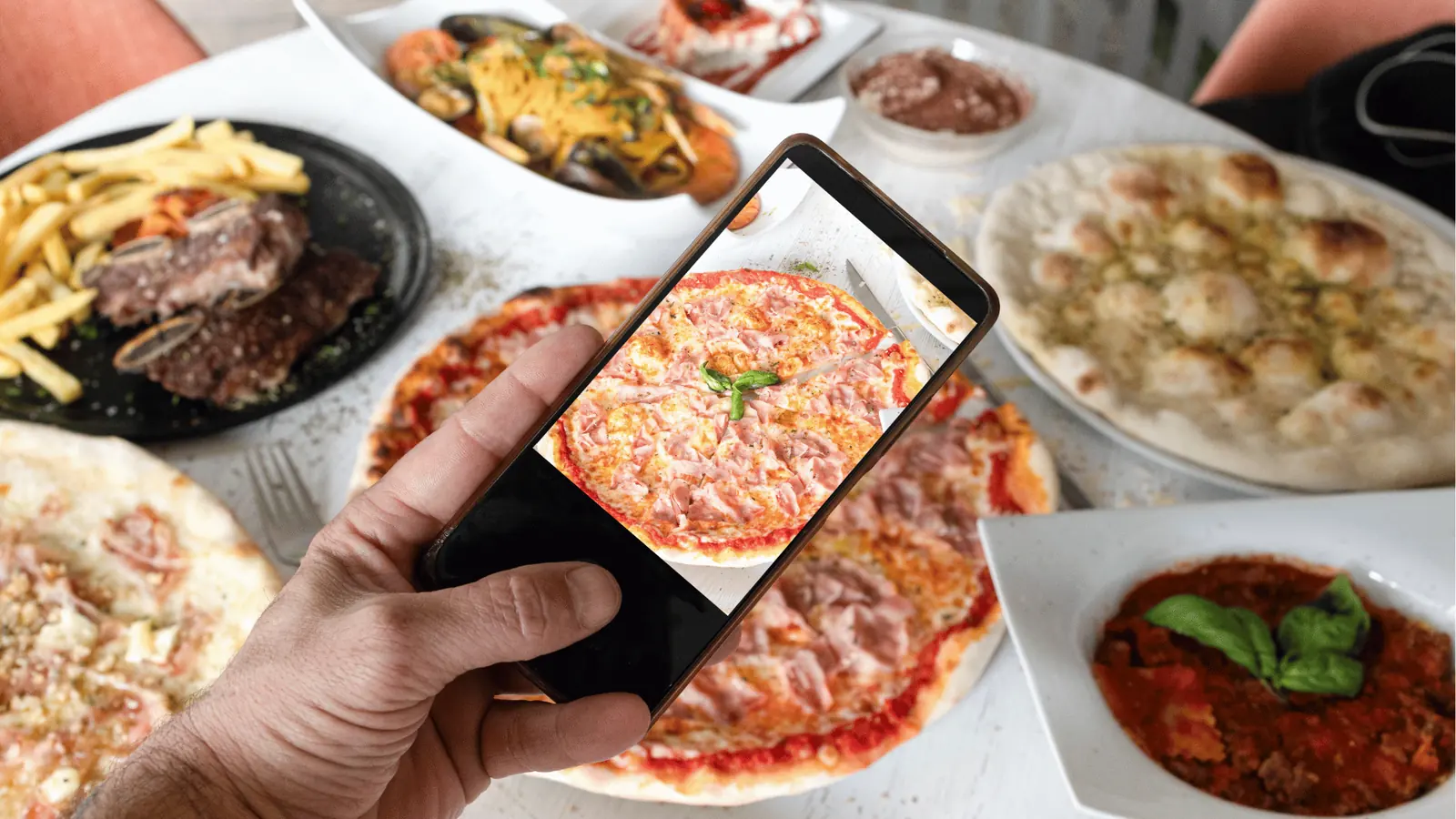 Hand holding phone photographing pizza on a table with multiple dishes