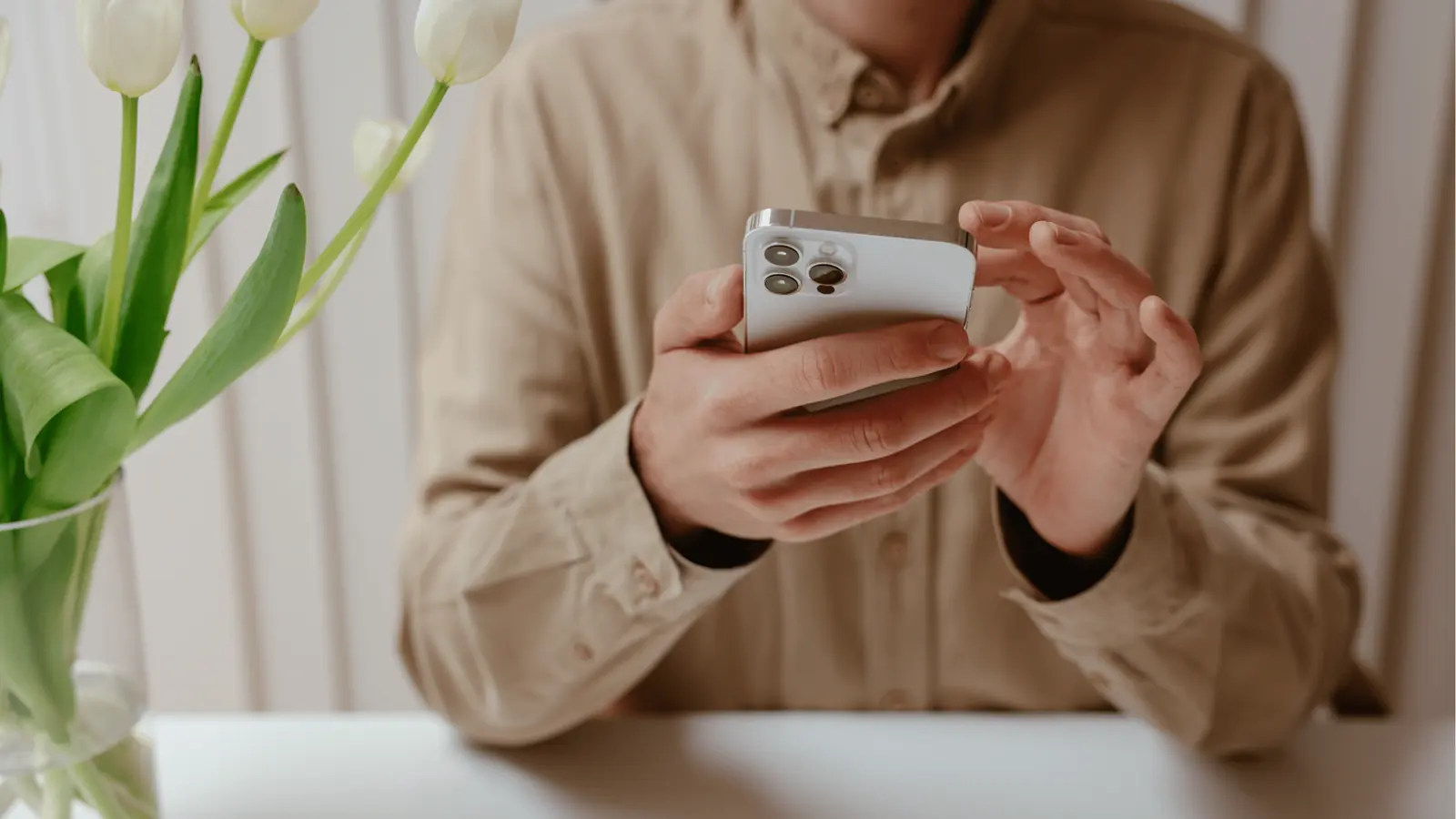 Person in beige shirt holding a smartphone at a white table with tulips