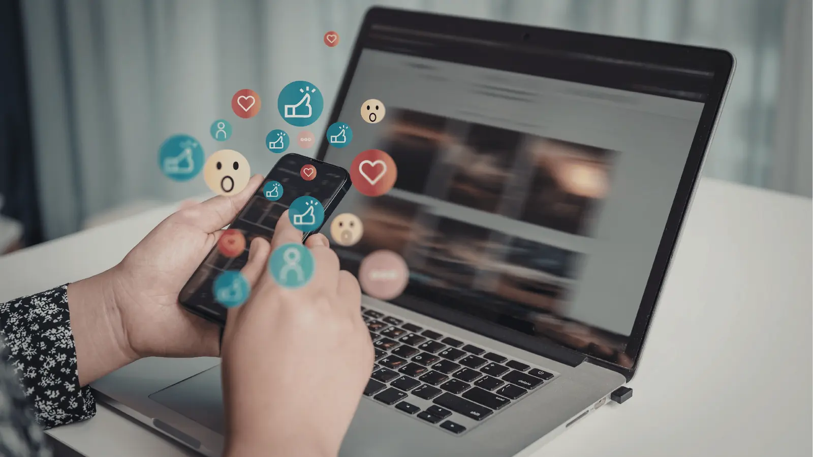 Hands holding smartphone over laptop with floating social reaction icons for publishing