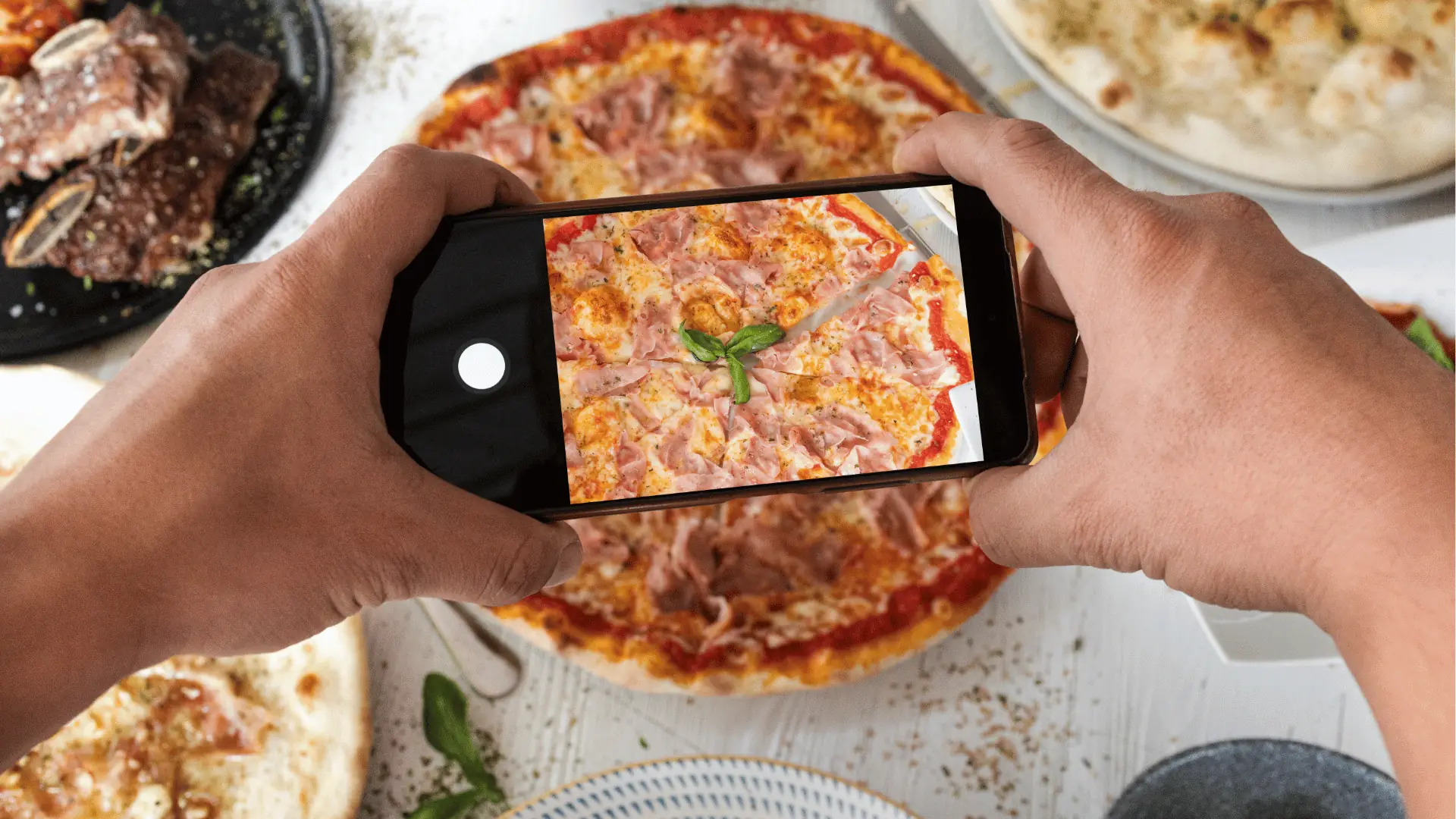 Two hands holding phone photographing a whole pizza on table