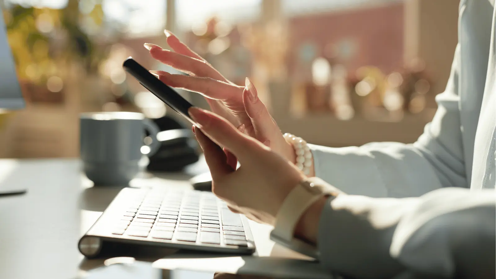 Person's hands using smartphone above keyboard on sunlit office desk