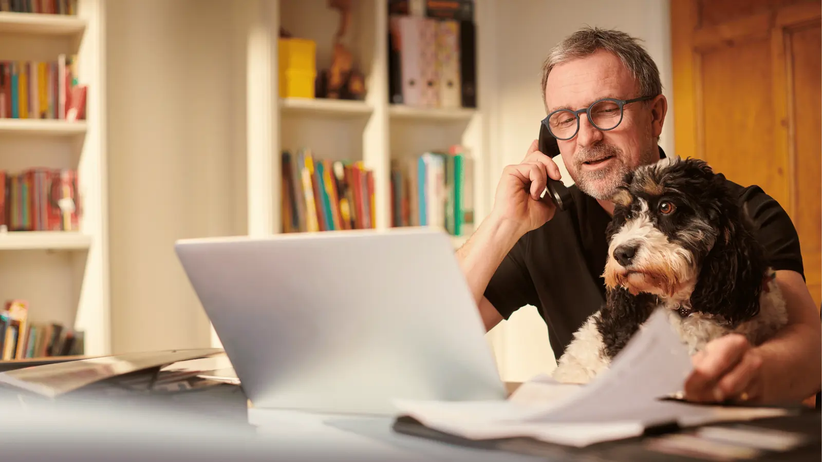 Middle aged man on phone at laptop with black and white dog