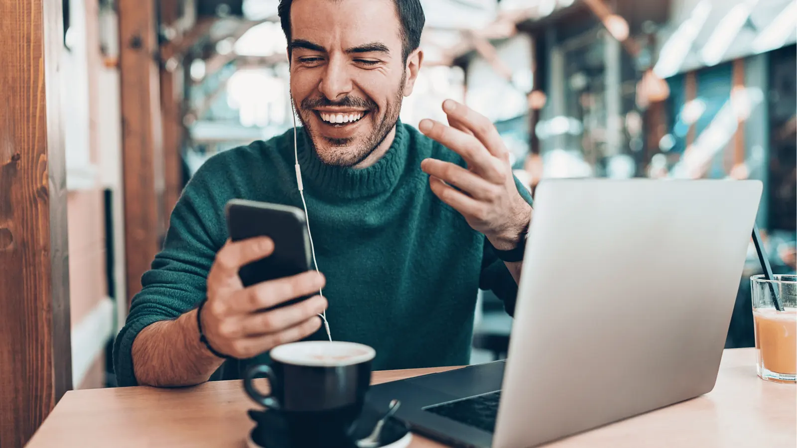 Smiling man in cafe using phone with laptop, coffee, and earphones