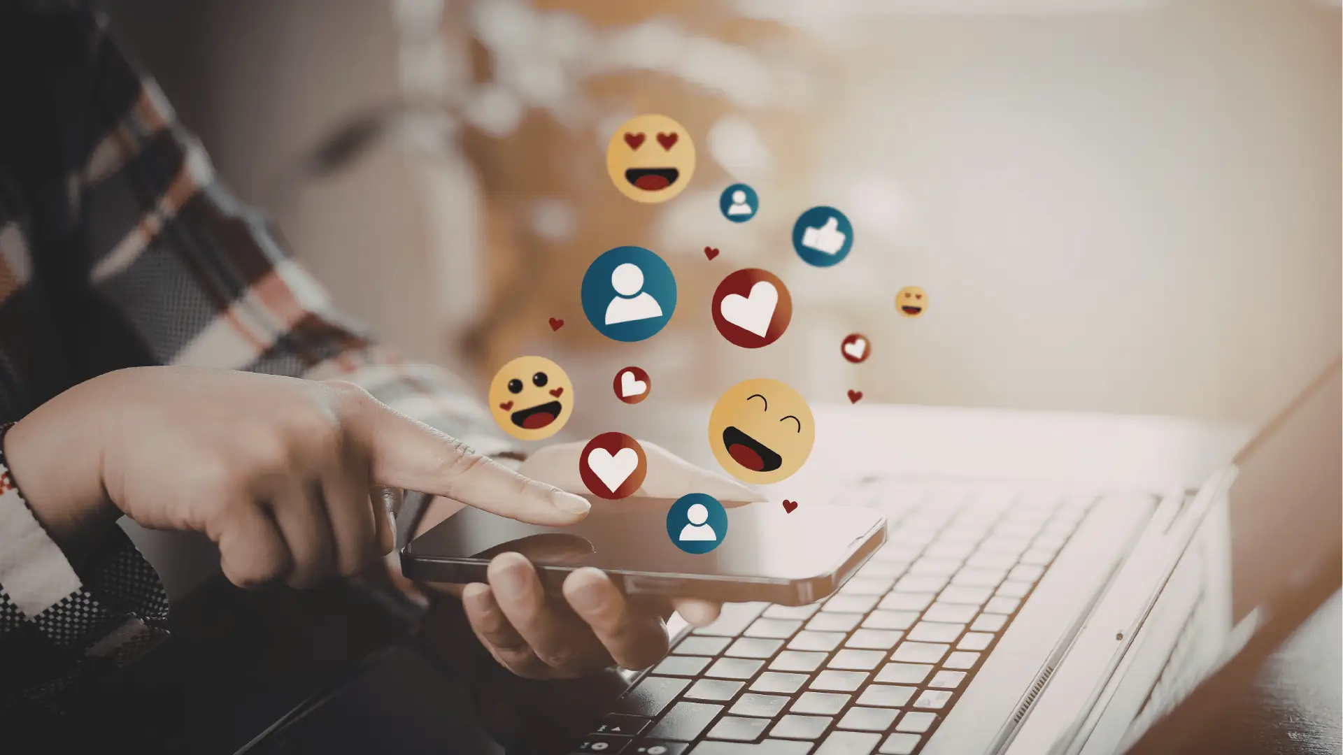 Hands tapping smartphone with floating social media reaction emojis over laptop