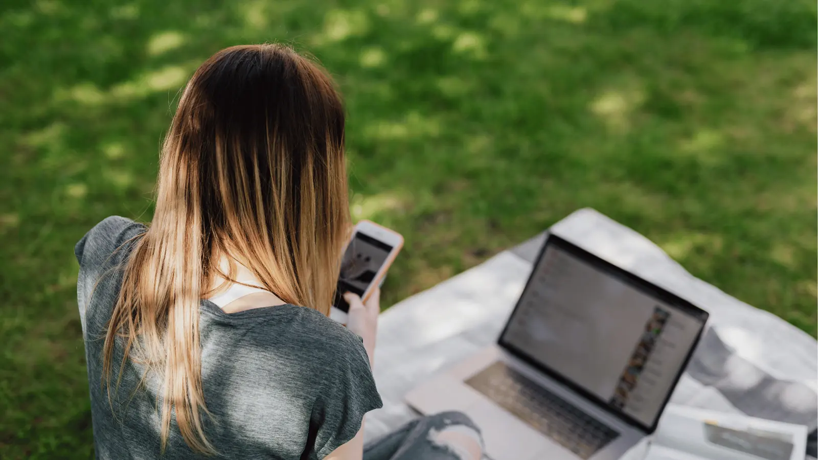 Person sitting on blanket outdoors using smartphone with laptop beside them