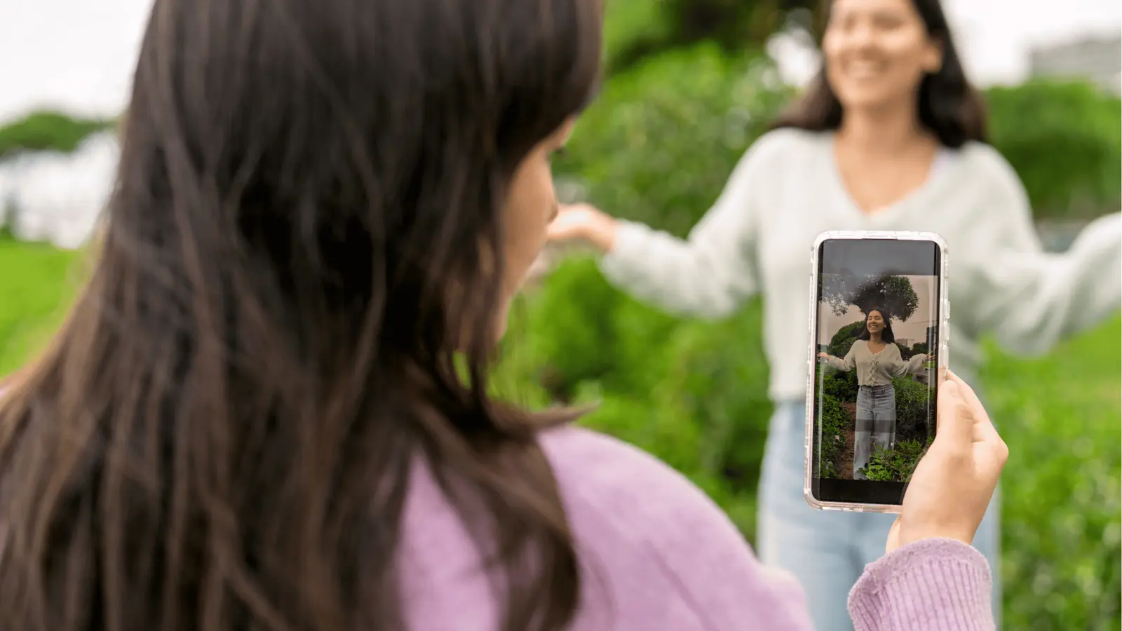 Person holding phone photographing friend posing with arms outstretched in park