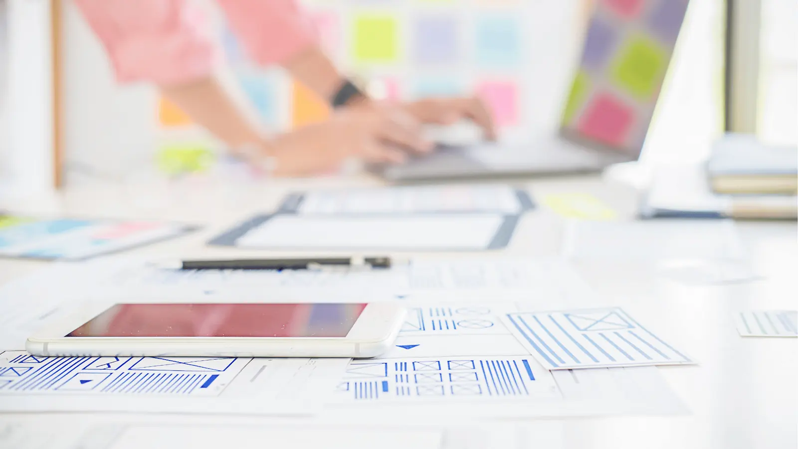 Smartphone rests on printed wireframes with blurred hands typing on laptop