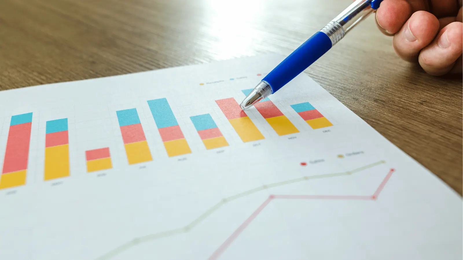 Hand holding a pen pointing at colorful stacked bar charts on paper