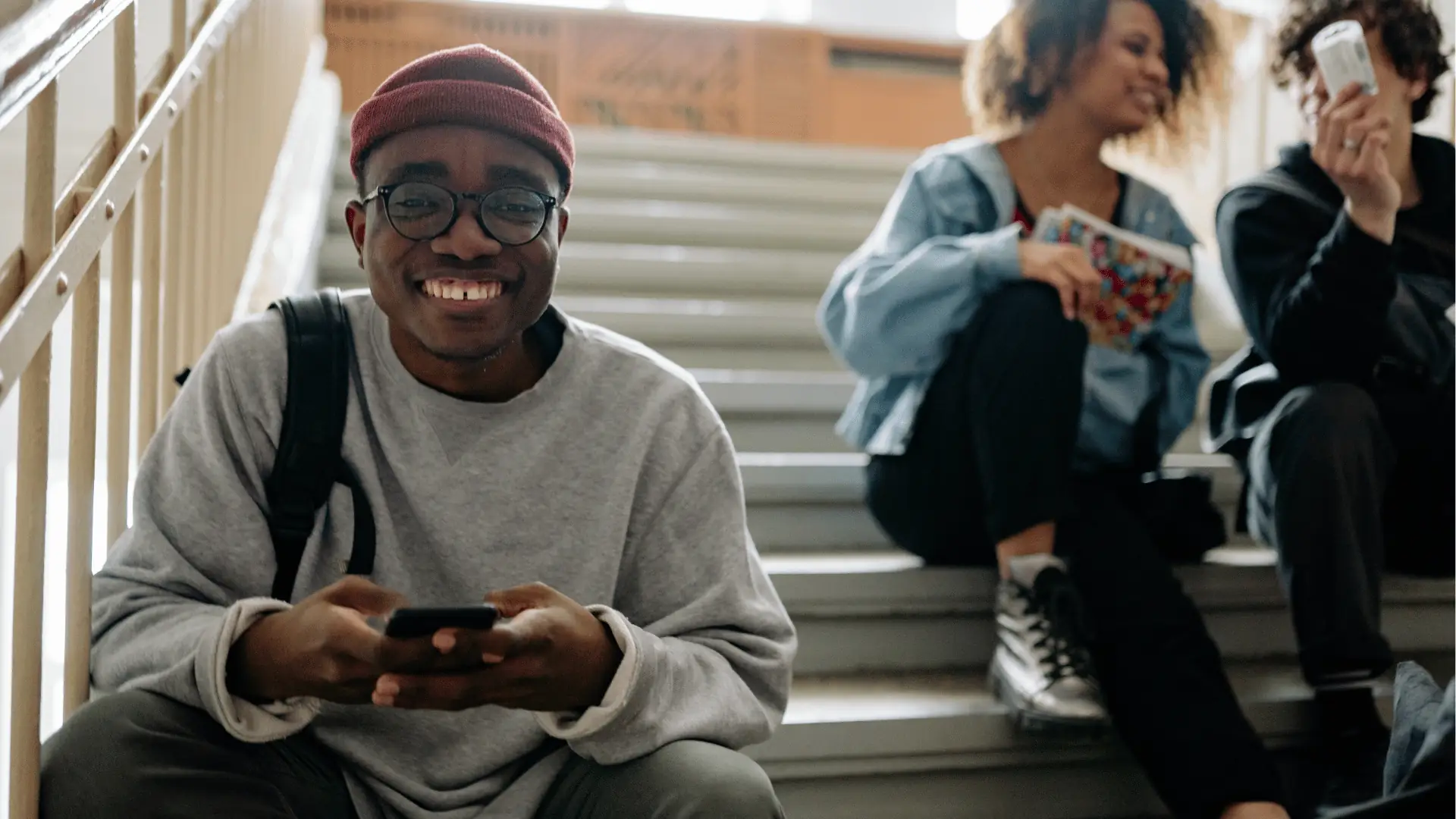 Smiling student with smartphone sitting on stairs with friends behind