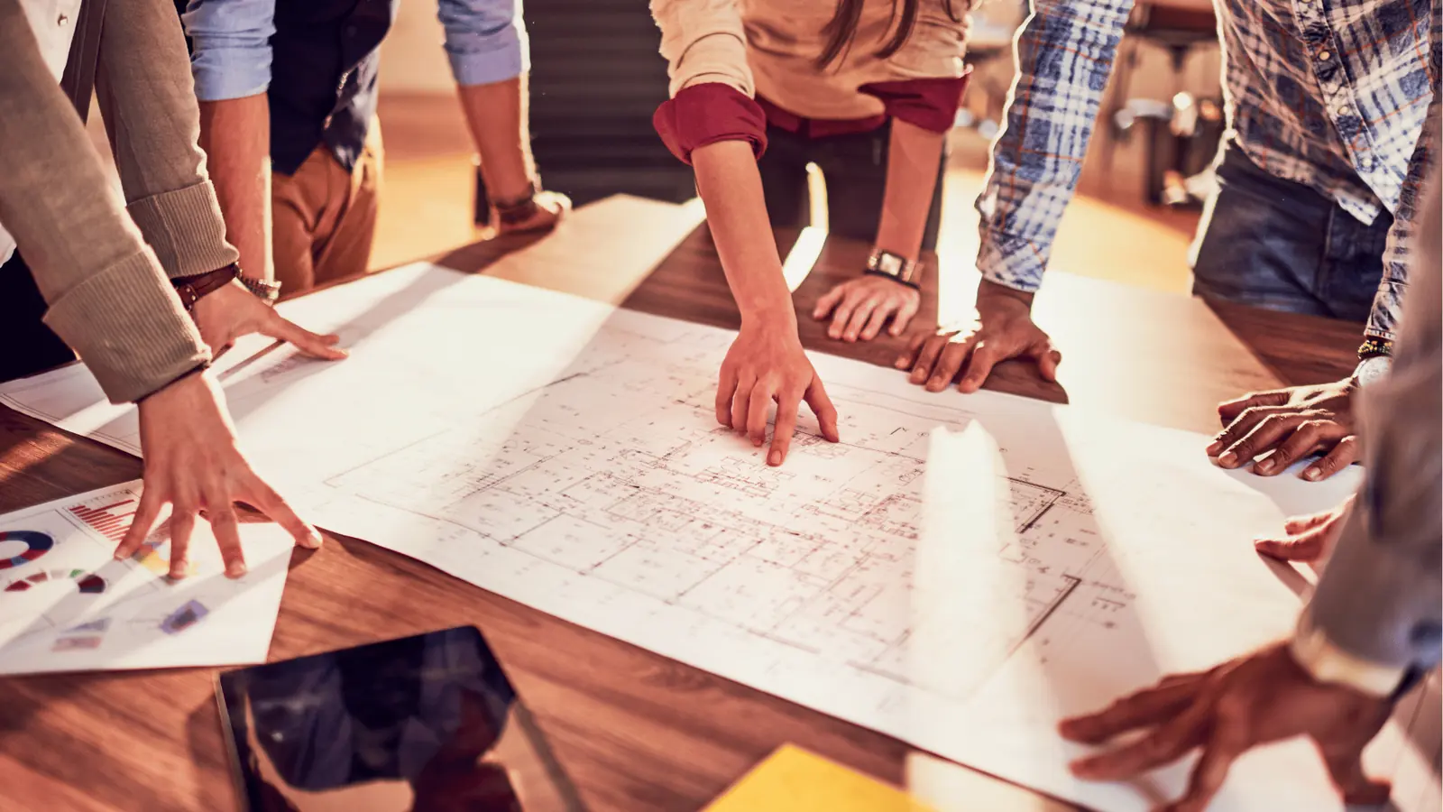 Team gathered around table pointing at large architectural blueprints and floor plans
