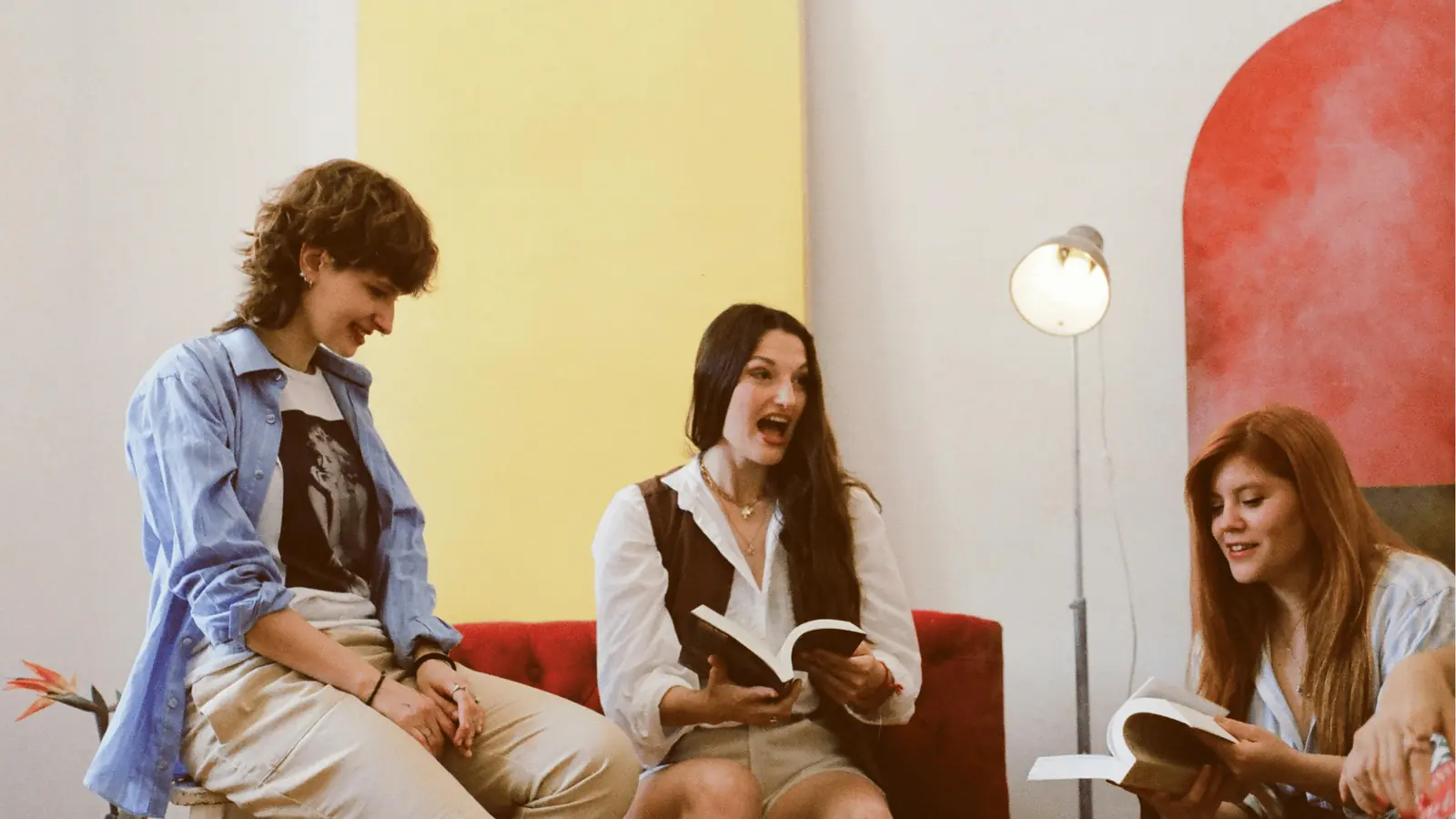 Three women sitting indoors reading books and animatedly talking on couch