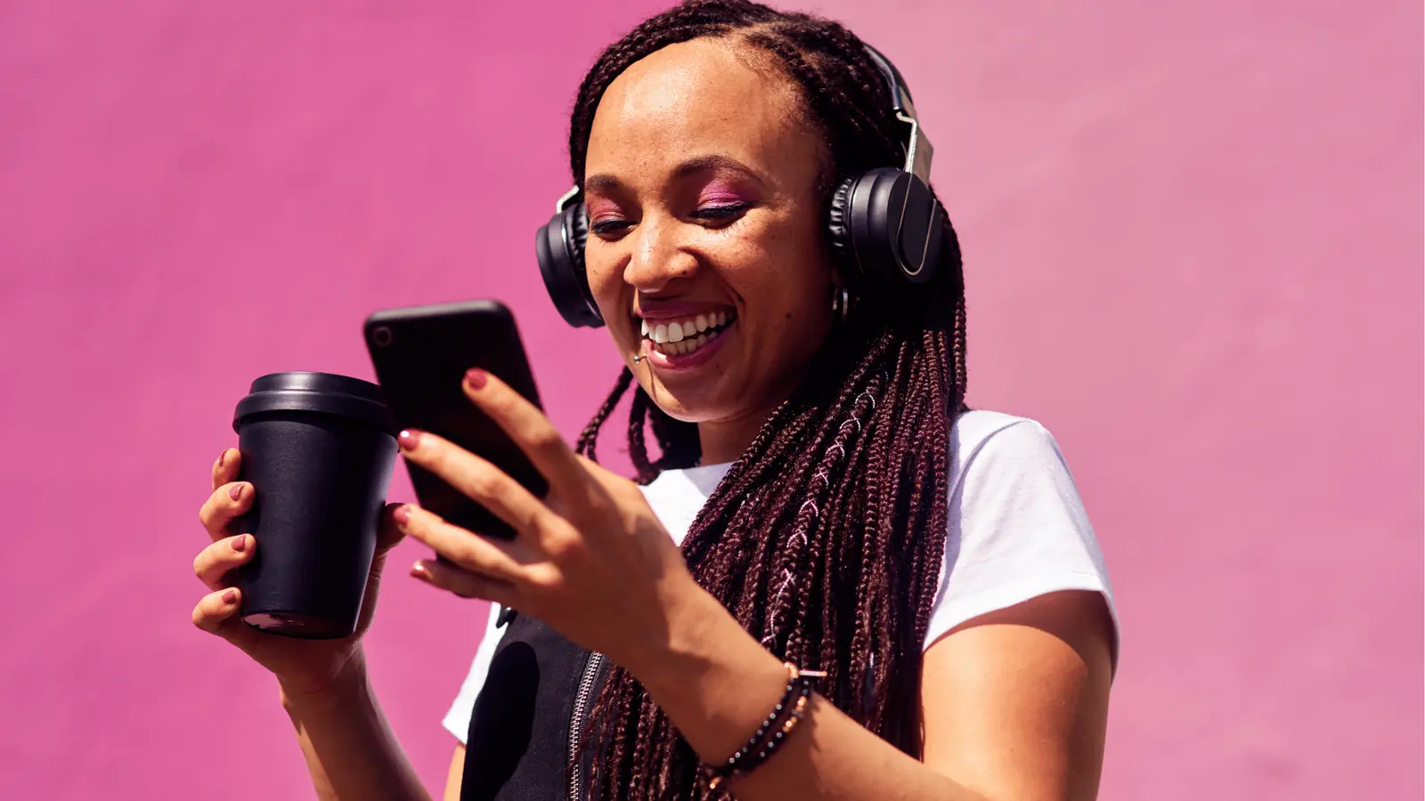 Smiling woman with headphones holding coffee cup and checking smartphone against pink wall