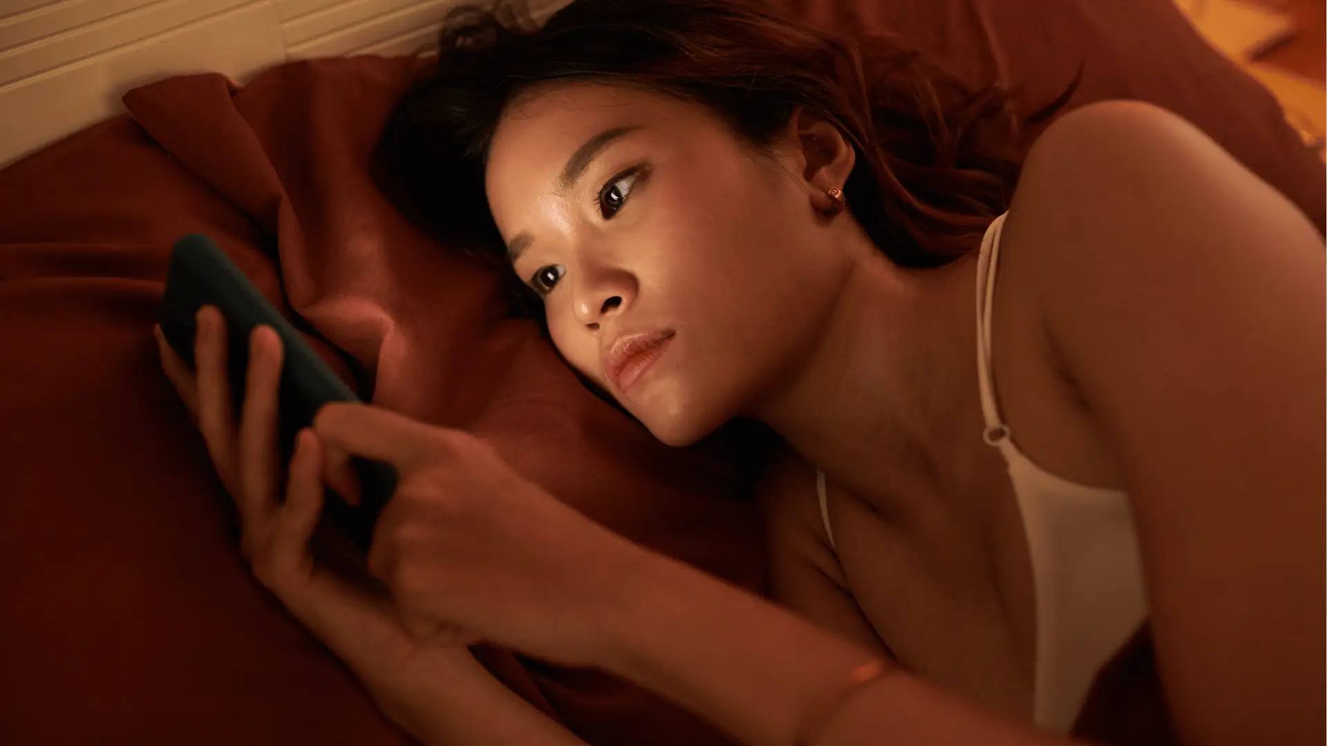 Young woman lying on bed looking intently at smartphone in warm light