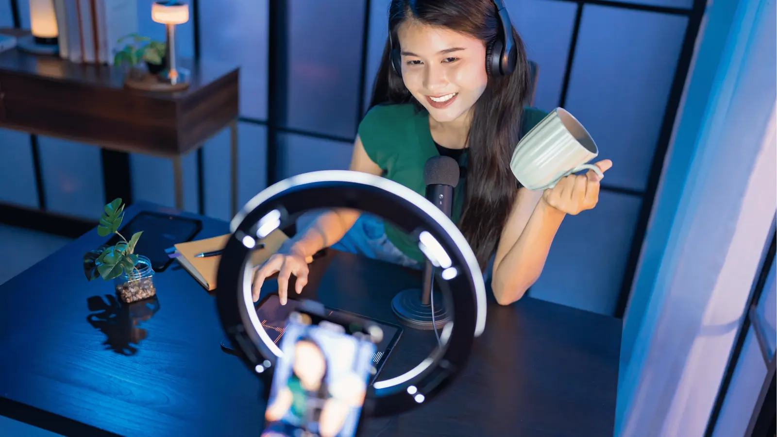 Smiling woman wearing headphones livestreaming at desk with mug and ring light for approval workflow