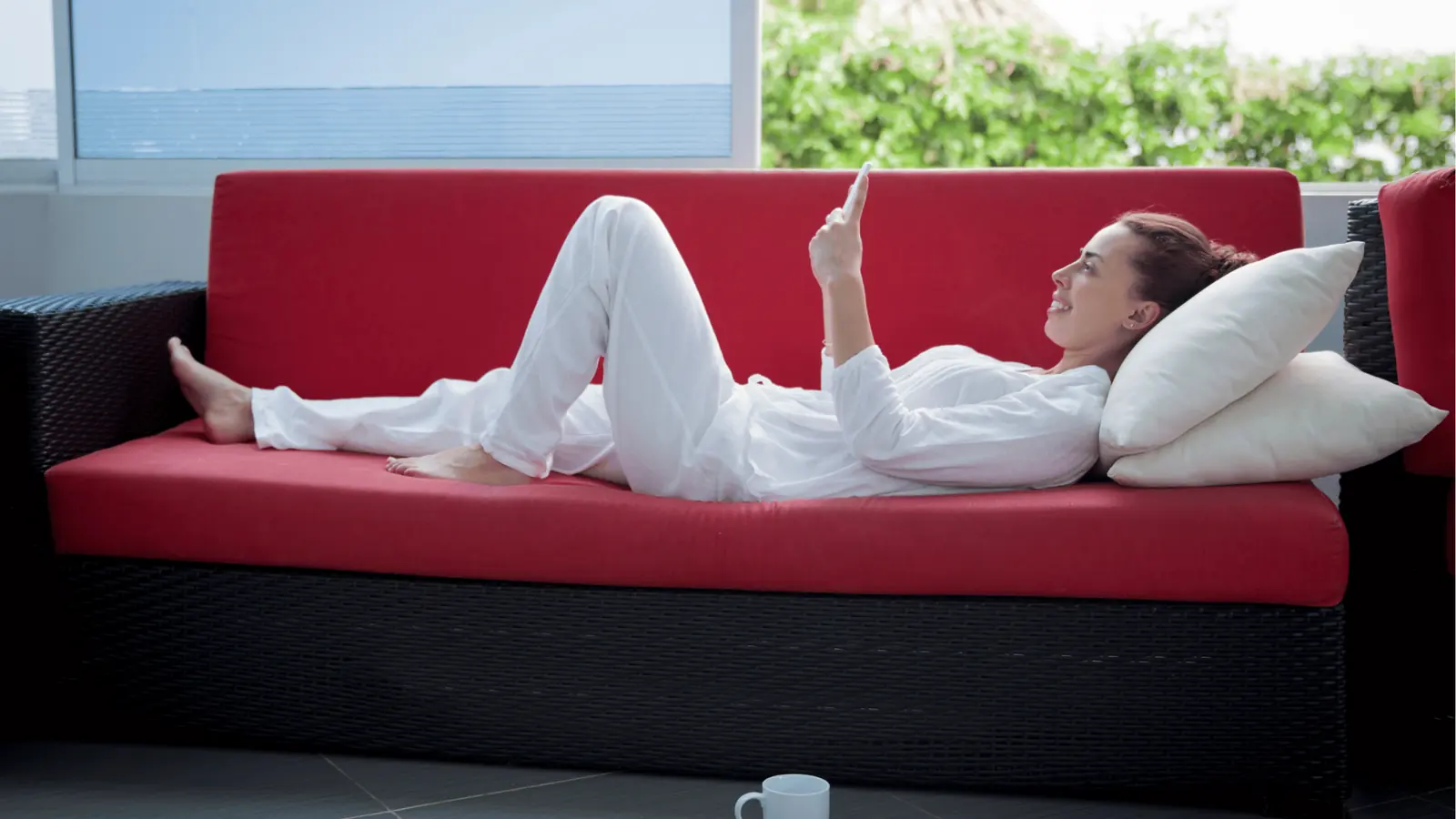 Woman lying on red sofa using smartphone with pillows and mug nearby