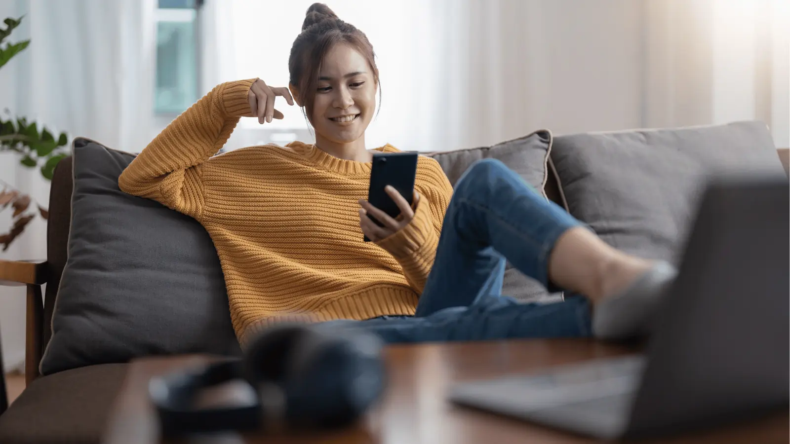 Young woman in yellow sweater reclining on couch and smiling at her phone