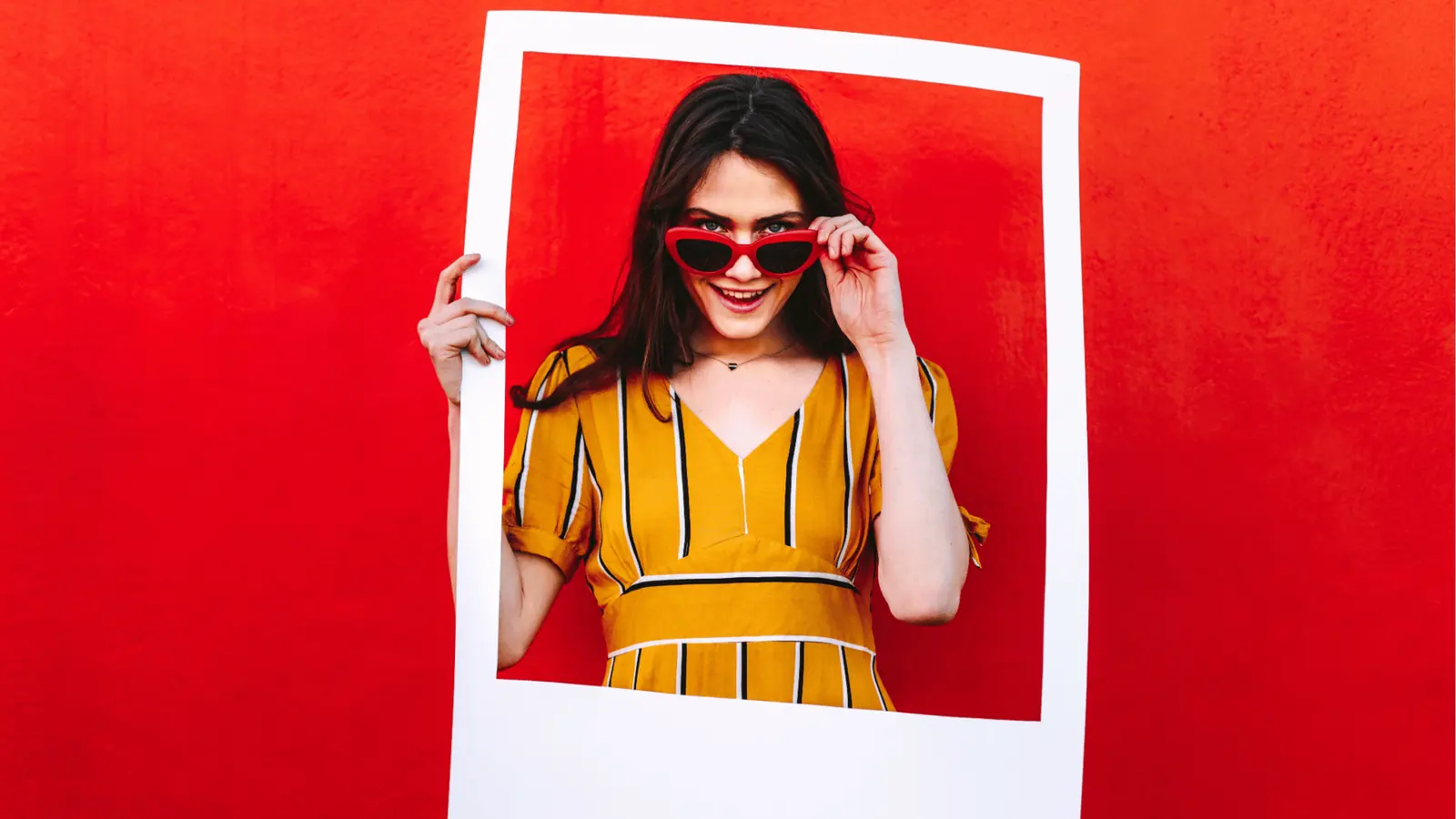 Young woman in yellow dress holds polaroid frame against red wall