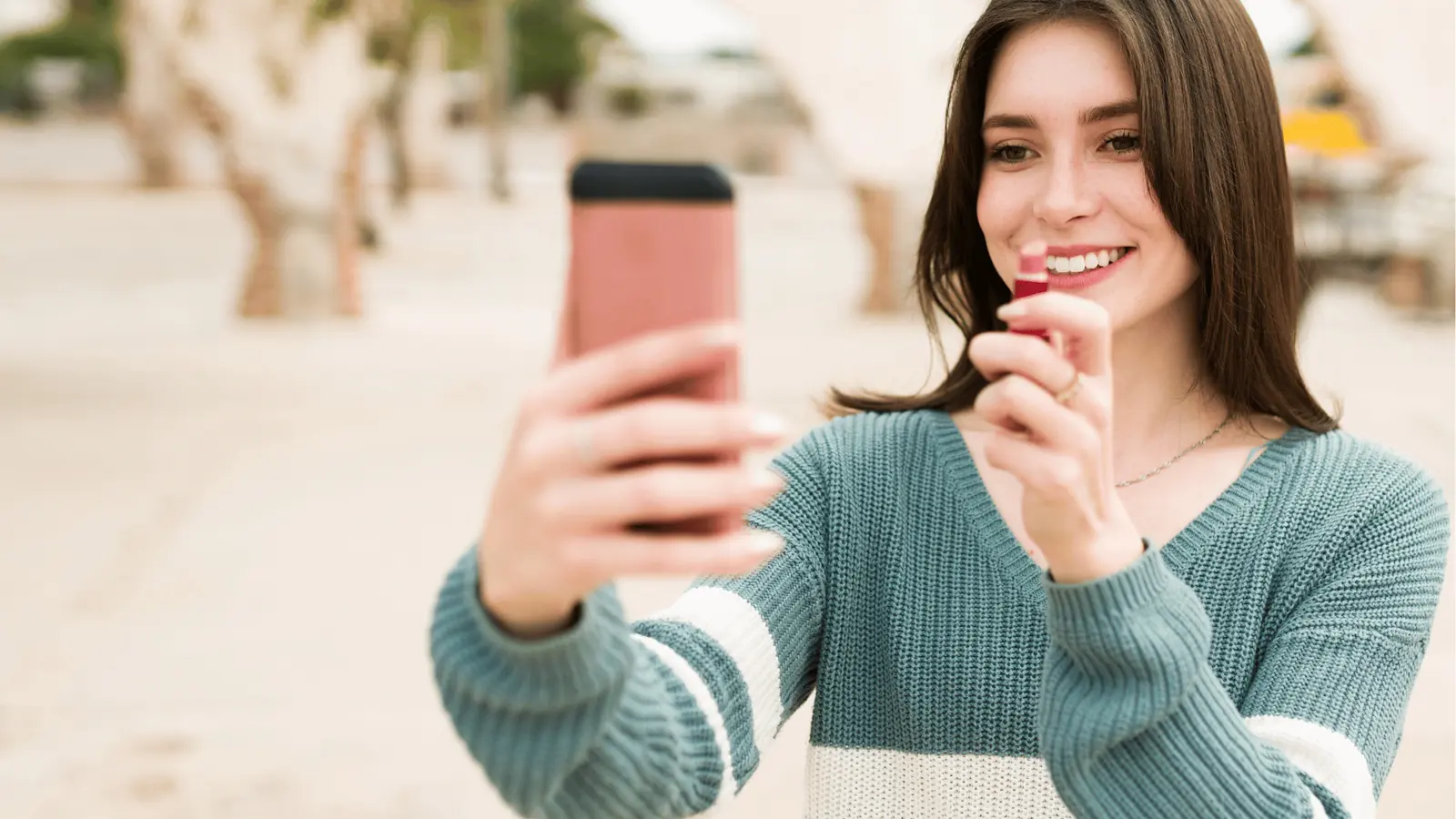 Young woman smiling while taking a lipstick selfie with smartphone outdoors for brand management
