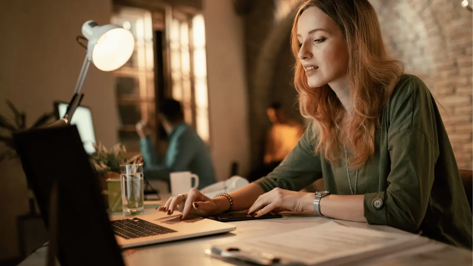 Woman typing on laptop at desk in warm coworking office with lamp