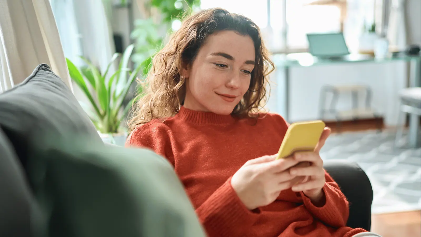 Woman in red sweater sitting on sofa looking at a yellow smartphone