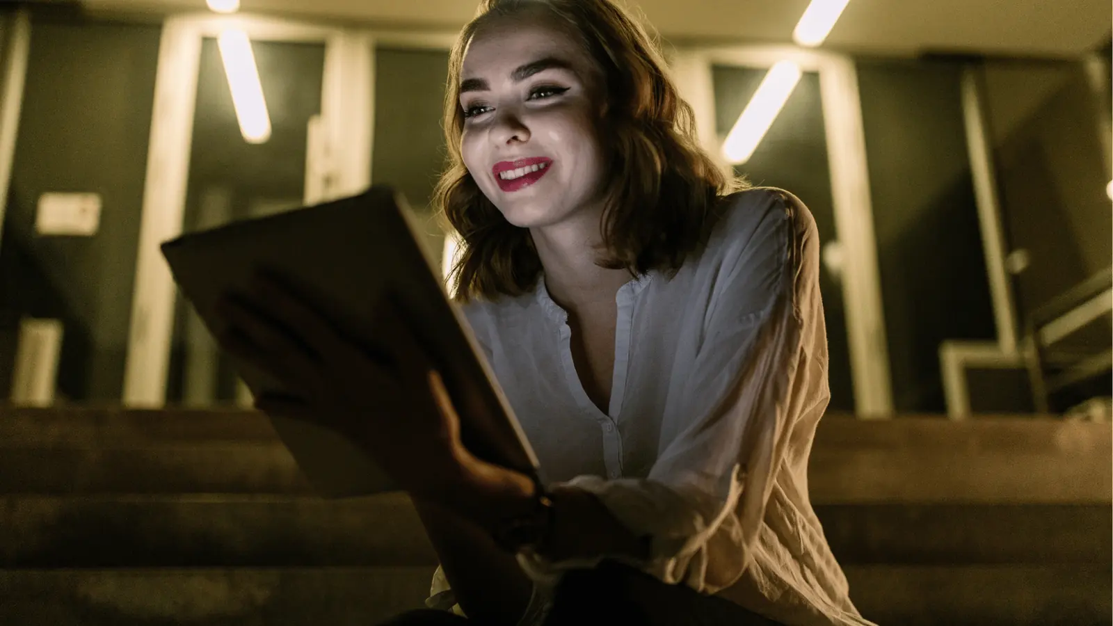 Young woman sitting on steps at night smiling while using a tablet