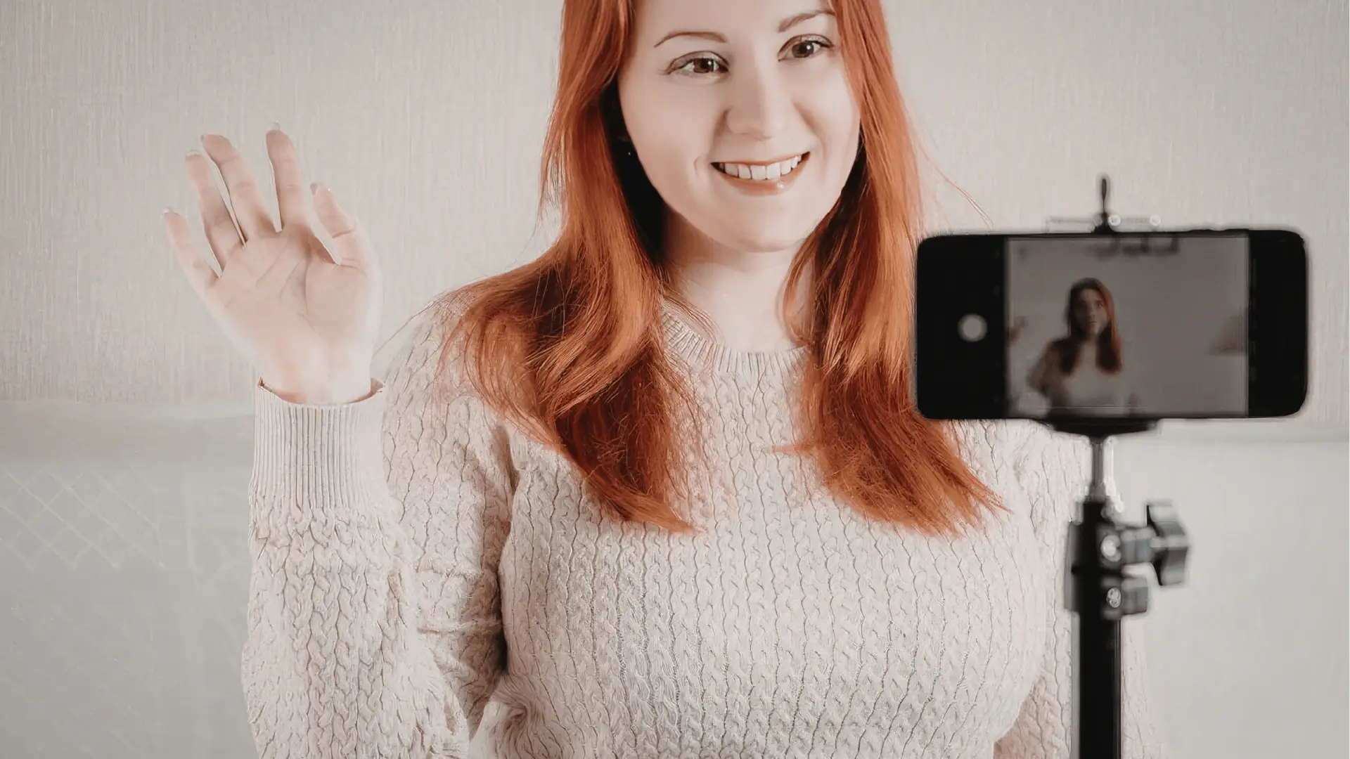 Smiling red-haired woman waving to smartphone camera on tripod for community management