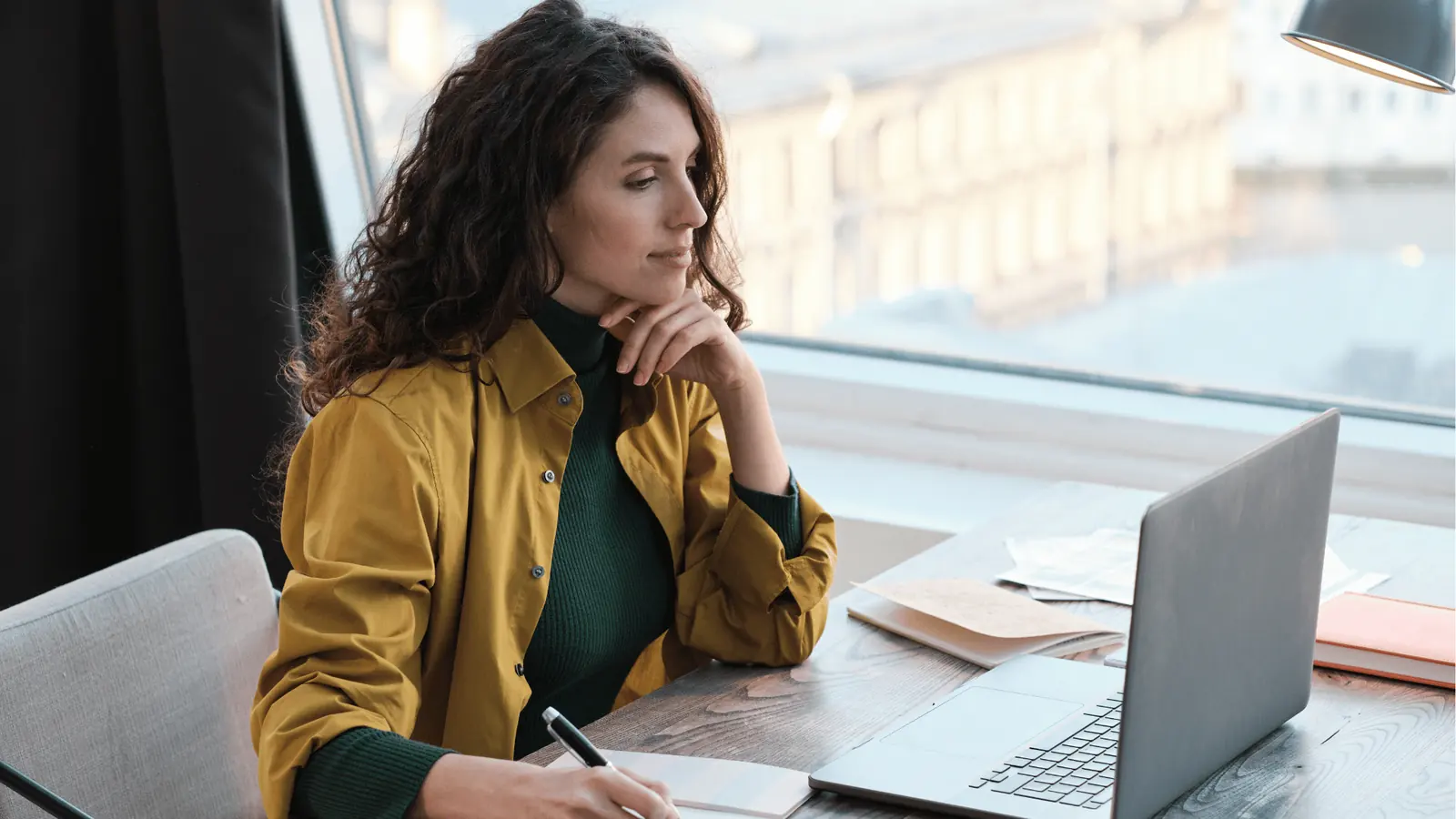 Woman in mustard jacket writing notes while looking at laptop by window for AI-assisted workflow