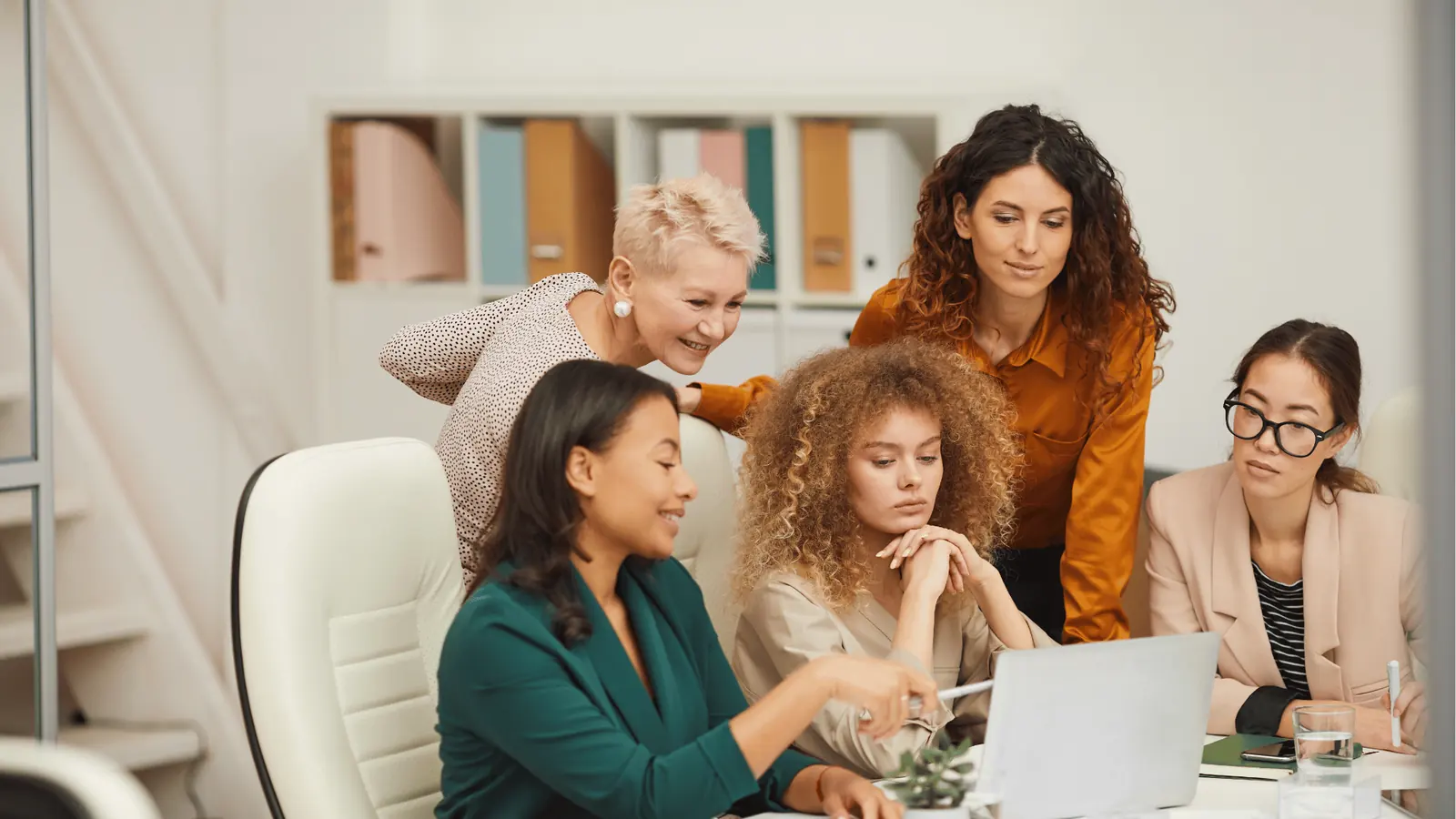 Five women gathered around a laptop in an office discussing work together