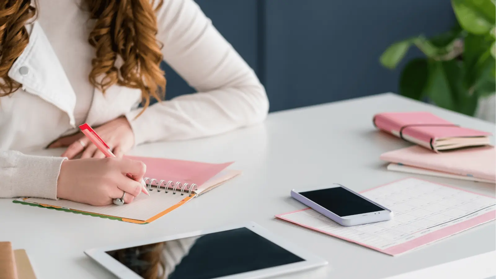 Person writing in a spiral planner at a white desk with phone