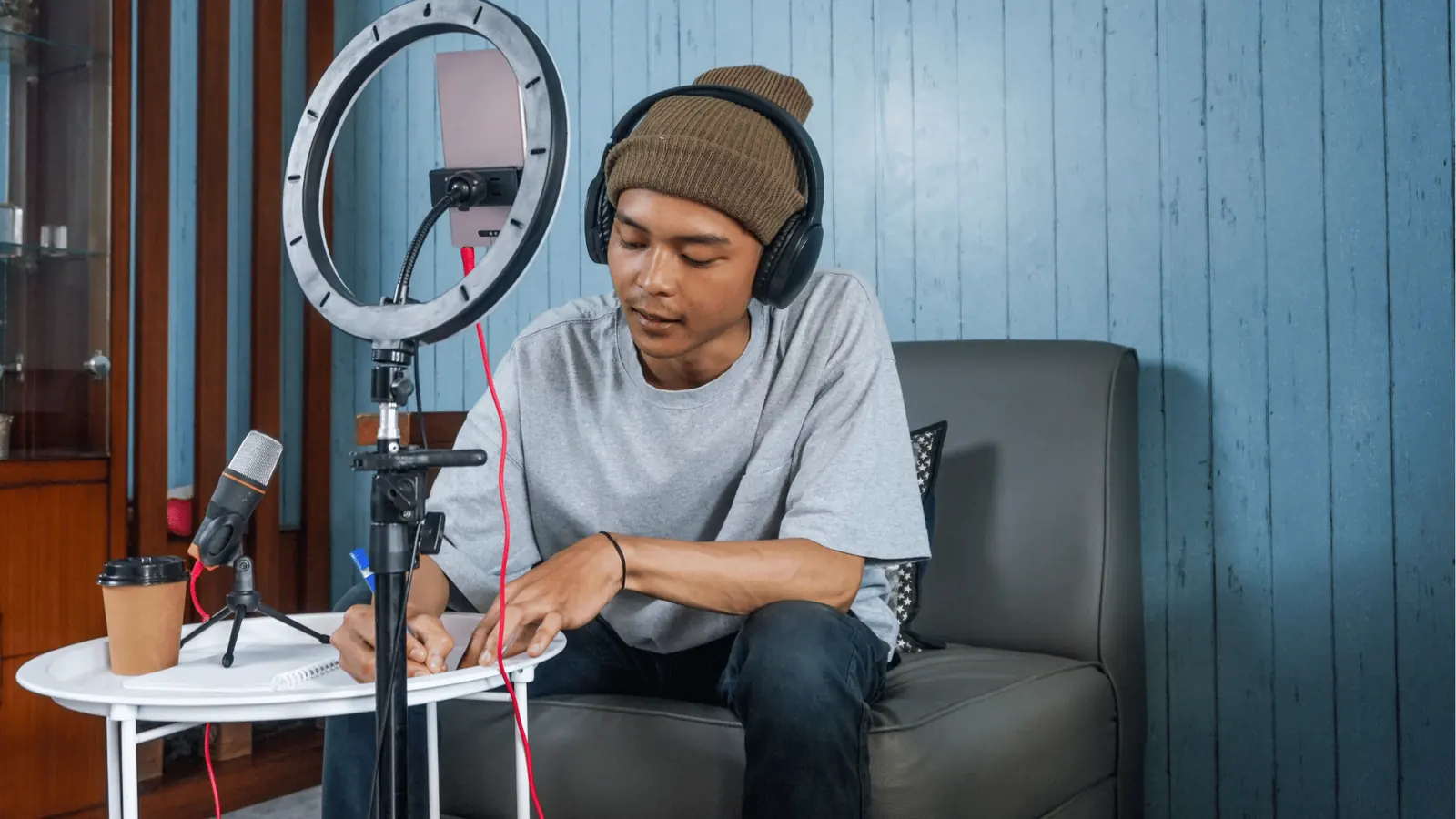 Young man with headphones writing notes while recording with ring light and microphone for reporting