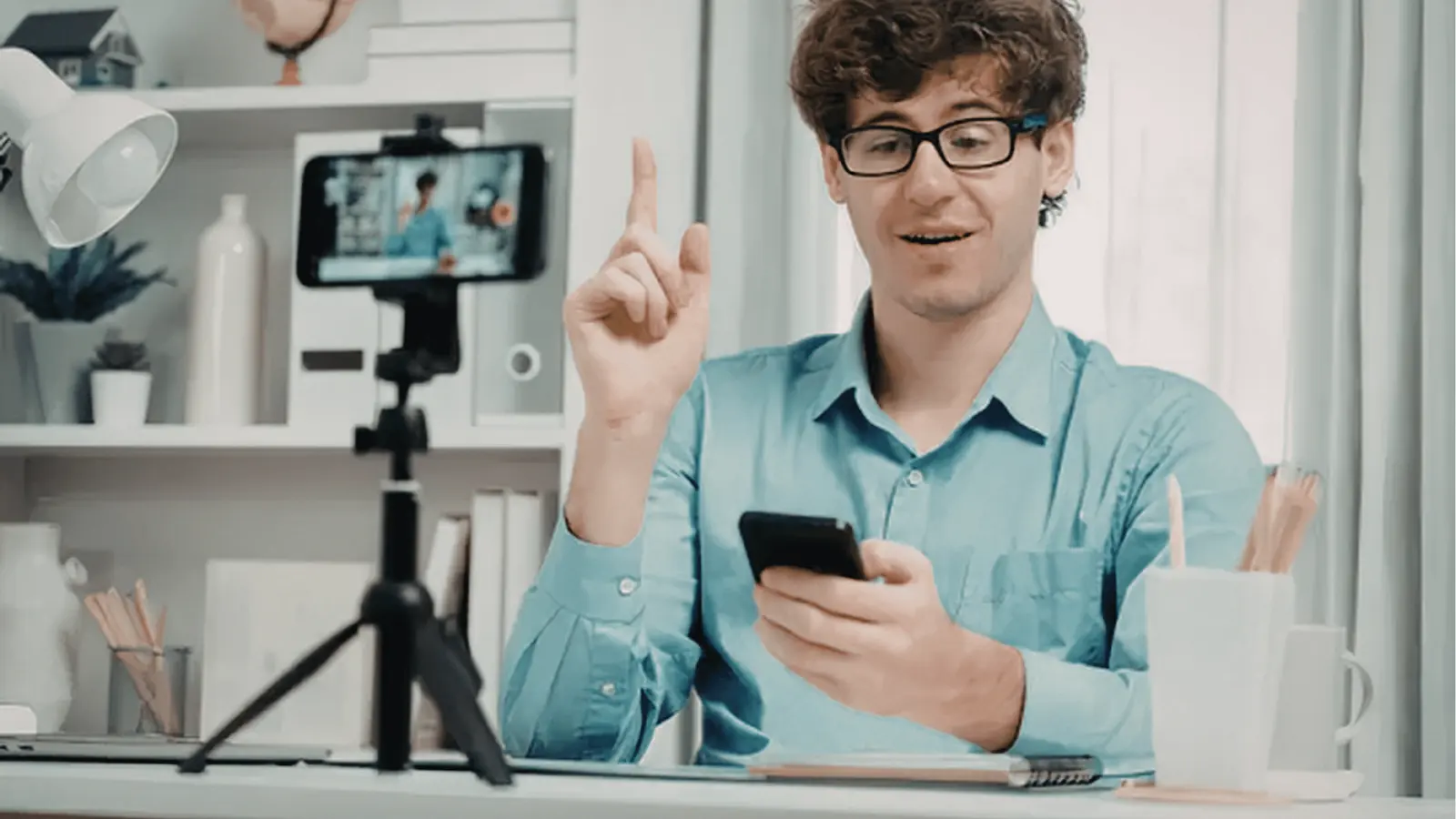 Young man in blue shirt gestures while recording with smartphone on tripod at desk
