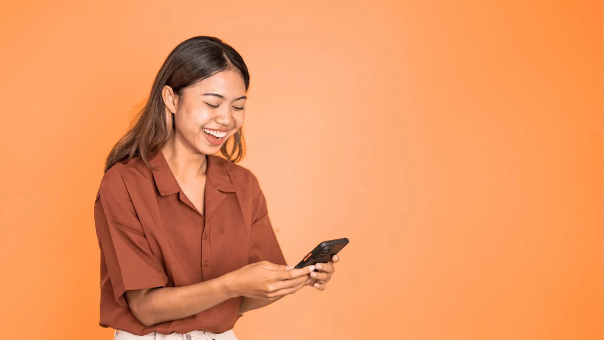 Young woman smiling while looking at smartphone against orange background